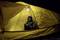 Een persoon zit 's nachts in een geel tent, draagt warme kleding en ziet er ontspannen uit. Een lamp verlicht het interieur van de tent.