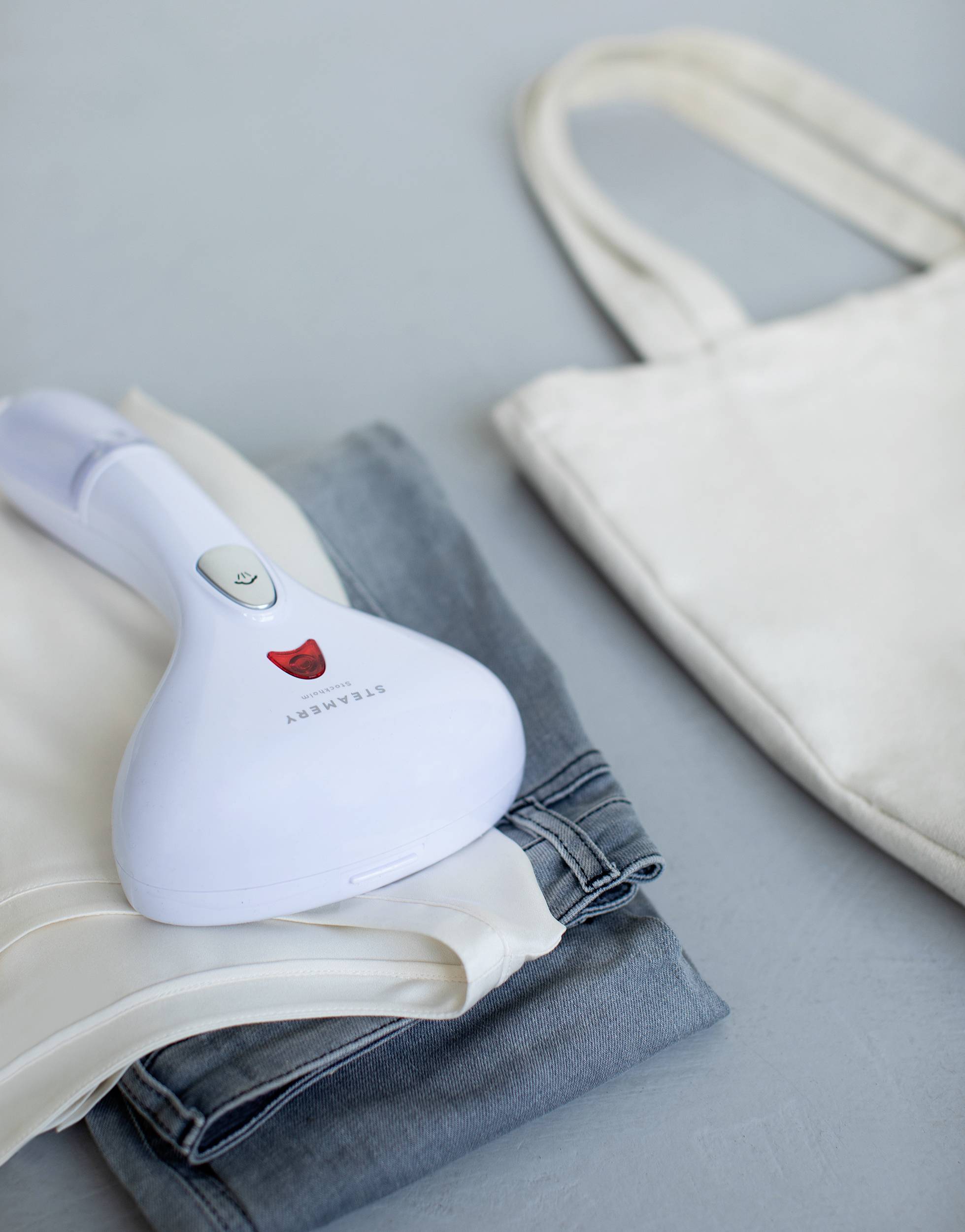 Een witte handstomer ligt gevouwen kleding naast een beige stoffen tas op een grijze achtergrond.
