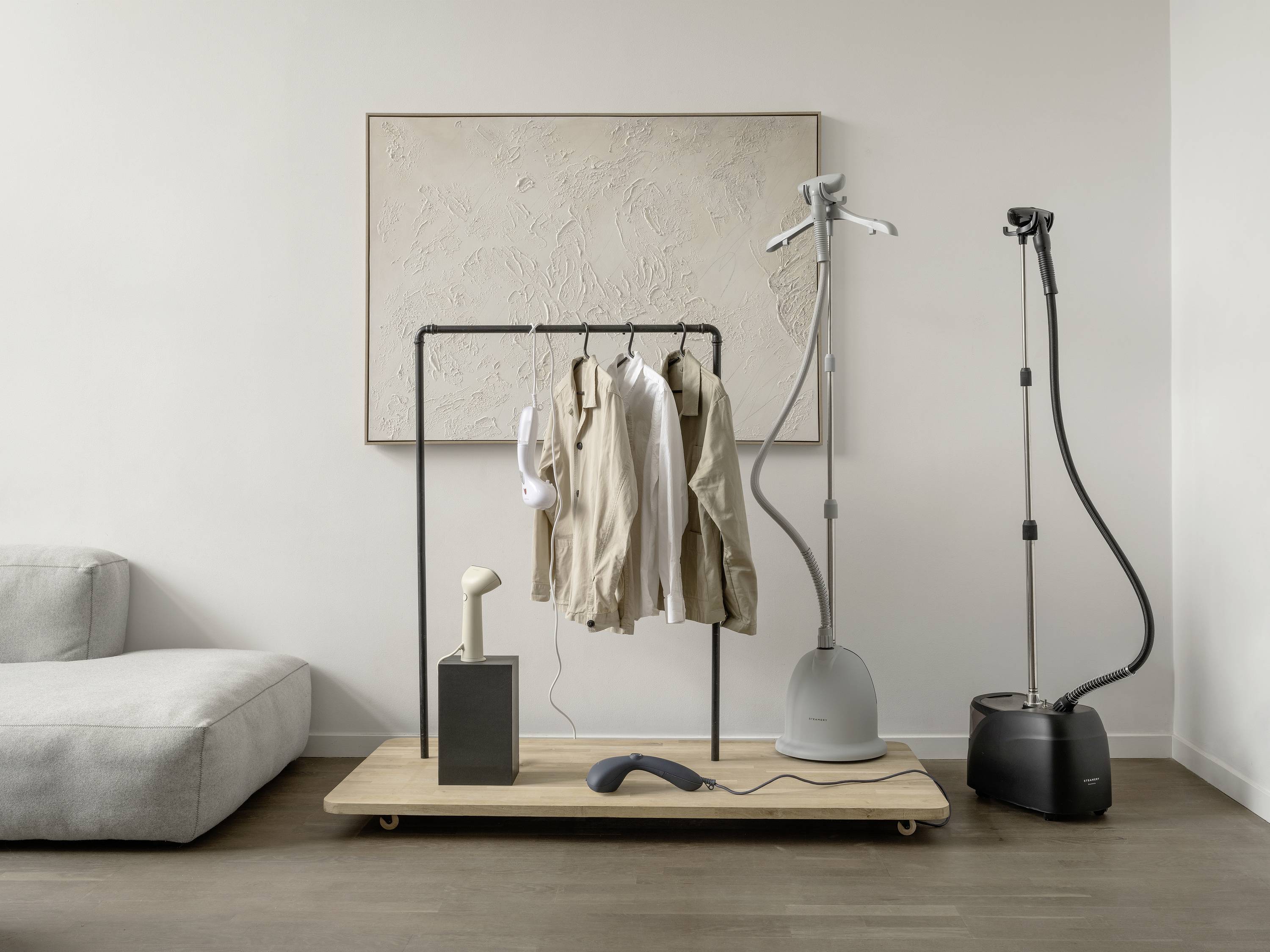 Een minimalistisch woonkamer toont een kledingrek met neutrale overhemden, een stoomstrijkijzer en een handstomer op een houten plank.