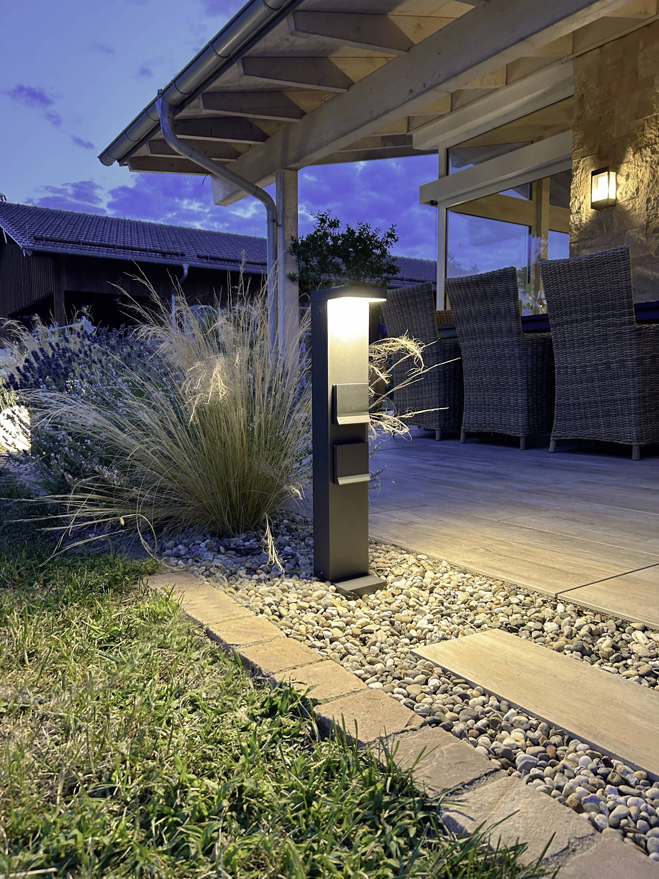 Terras bij schemering met moderne buitenverlichting, weilanden en grindpad. In de buurt staan tuinstoelen en een houten overkapping zorgt voor beschutting.