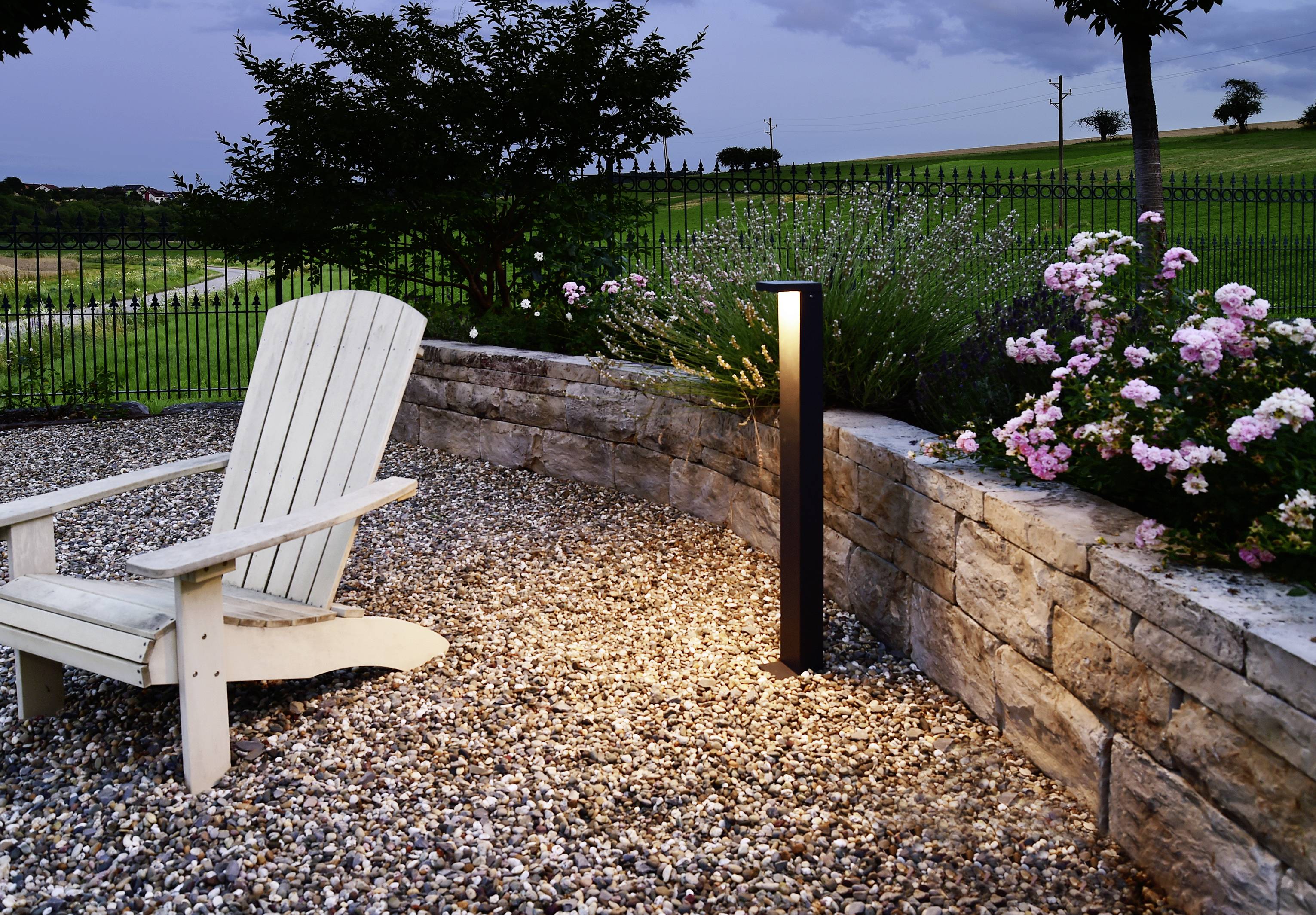 Een tuin met een stenen muur, verlicht door een staande lamp. Een witte houten stoel staat op een grindpad, omgeven door bloemen.