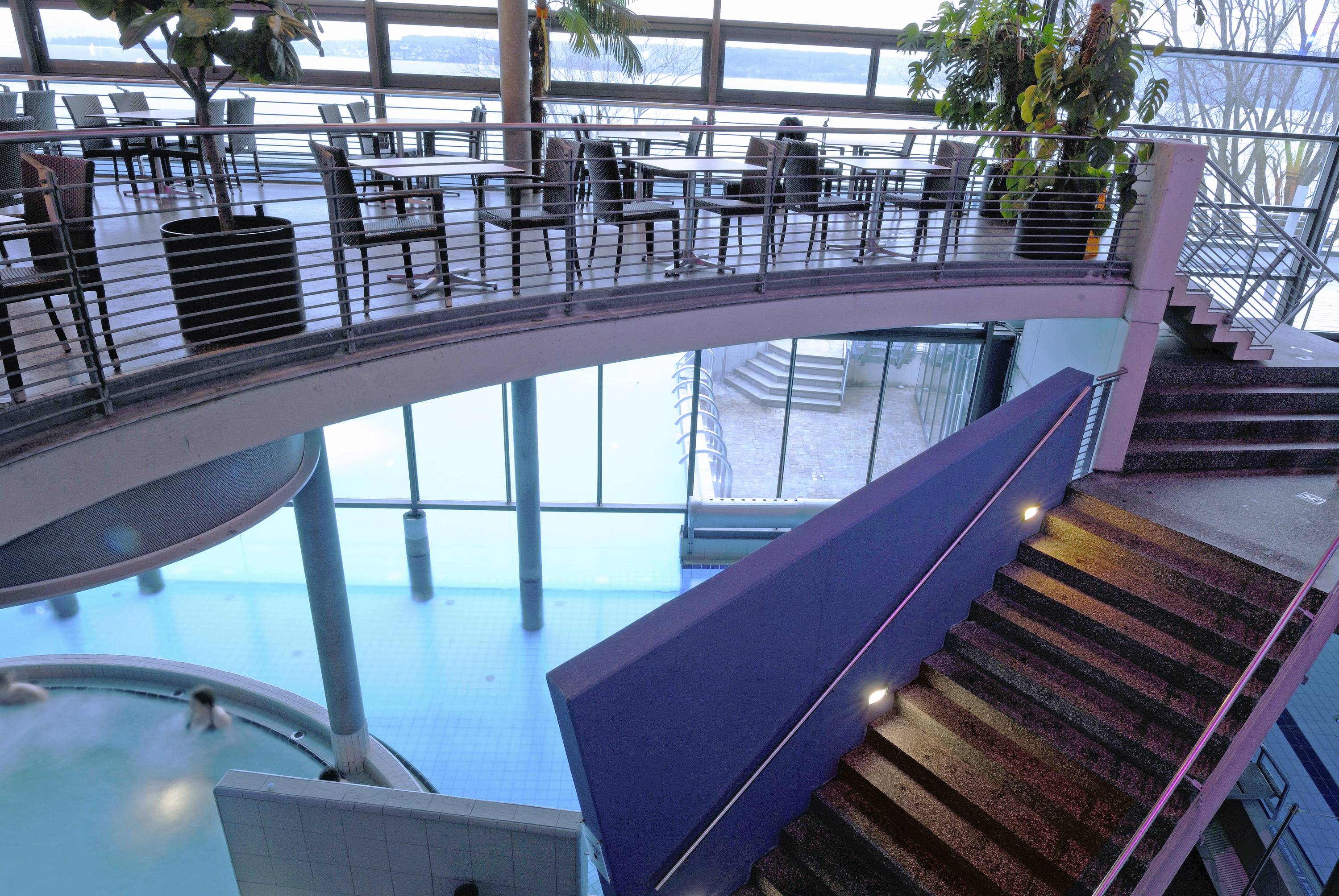 Binnenaanzicht van een modern zwembad met blauwe verlichting. Een trap leidt naar een verhoogd cafégedeelte met tafels en planten.