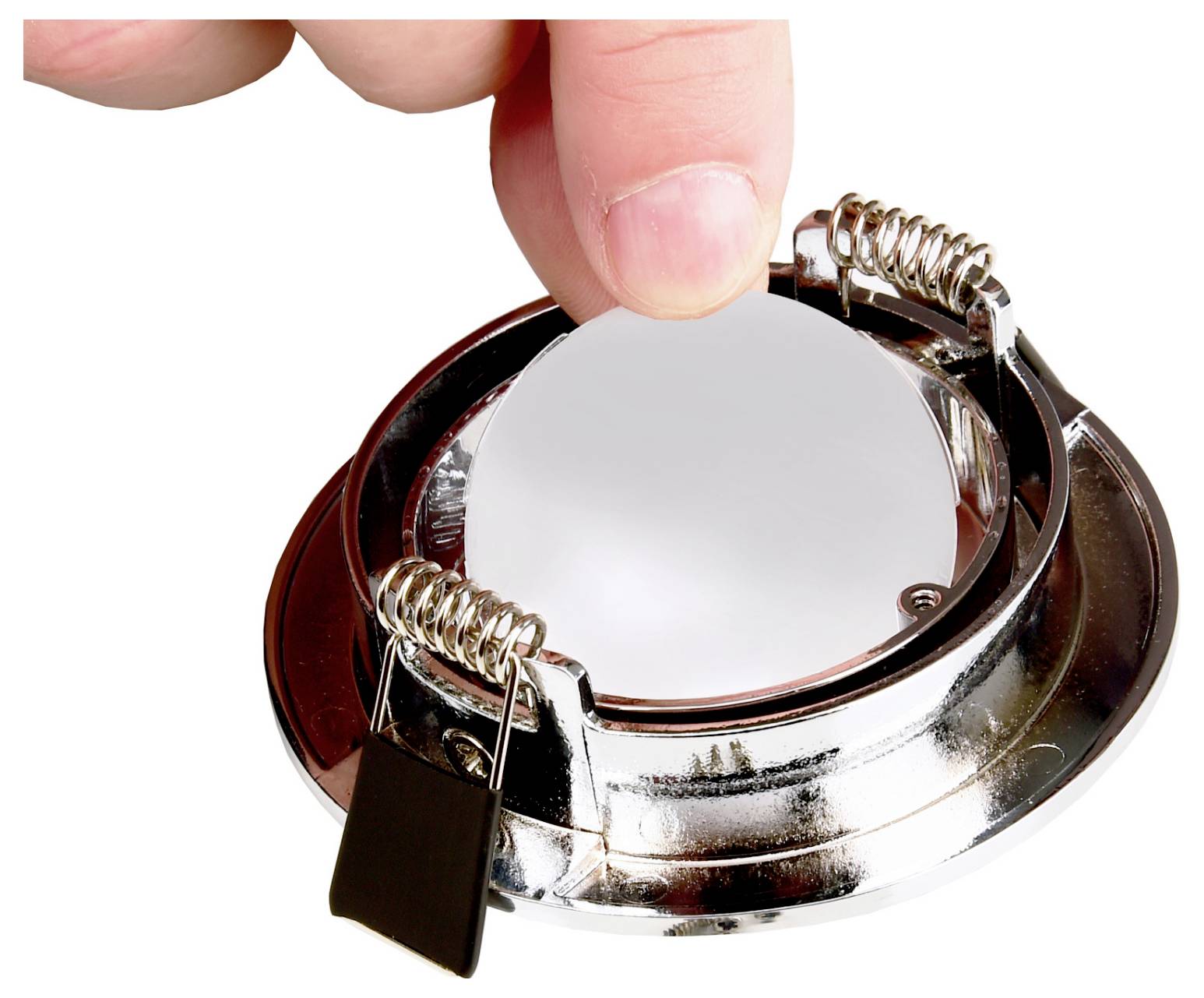 Een hand klemt een verzonken plafondlamp van chroom met veerklemmen vast tegen een witte achtergrond.