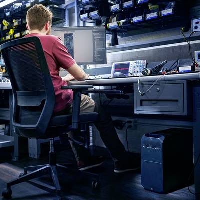 Een man zit aan een bureau en kijkt geconcentreerd naar computerbeeldschermen met code. Op tafel staan verschillende technologische apparaten.