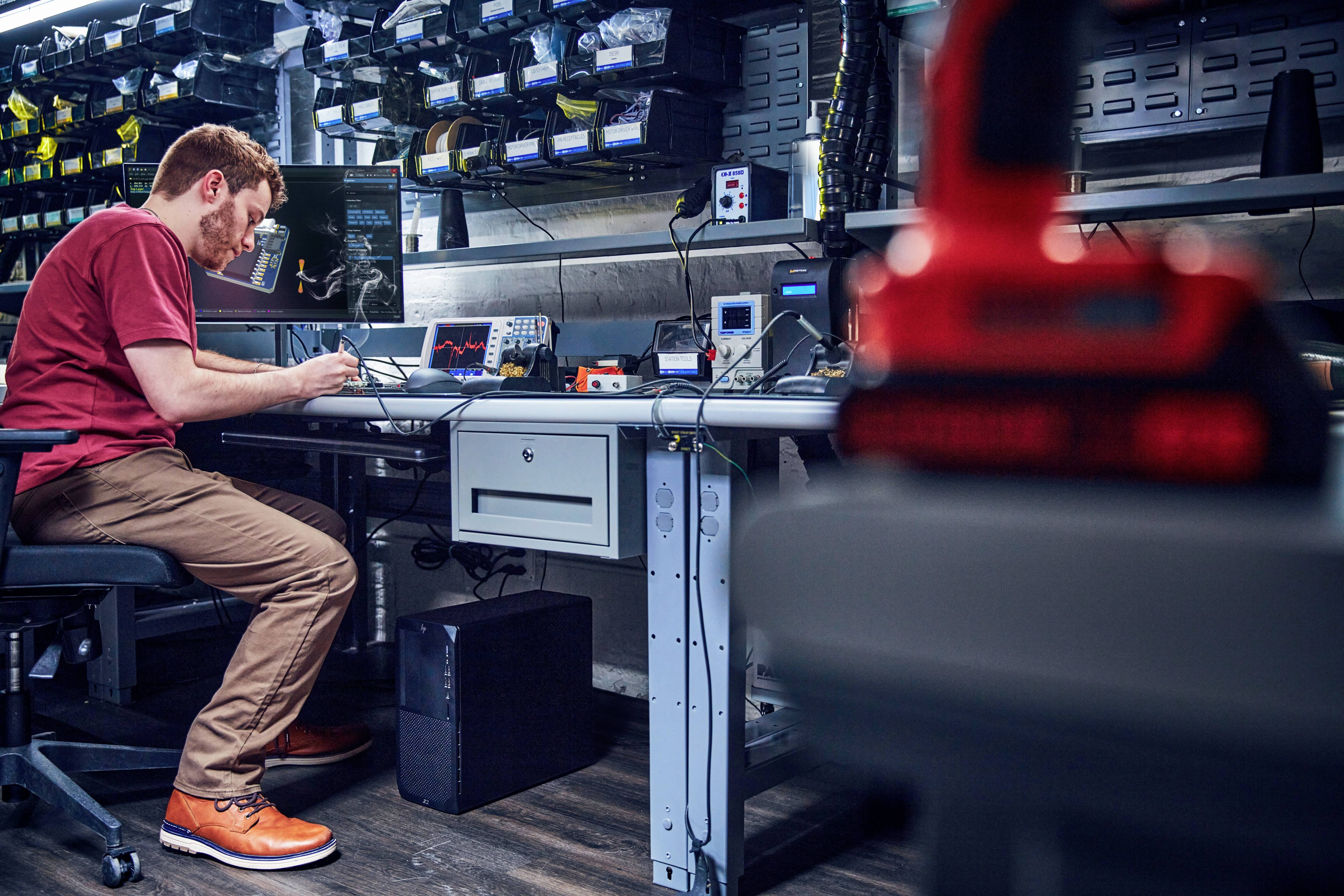 Een man werkt aan een laboratoriumtafel met elektronische apparaten en testapparatuur, omringd door schappen met kleine onderdelen.