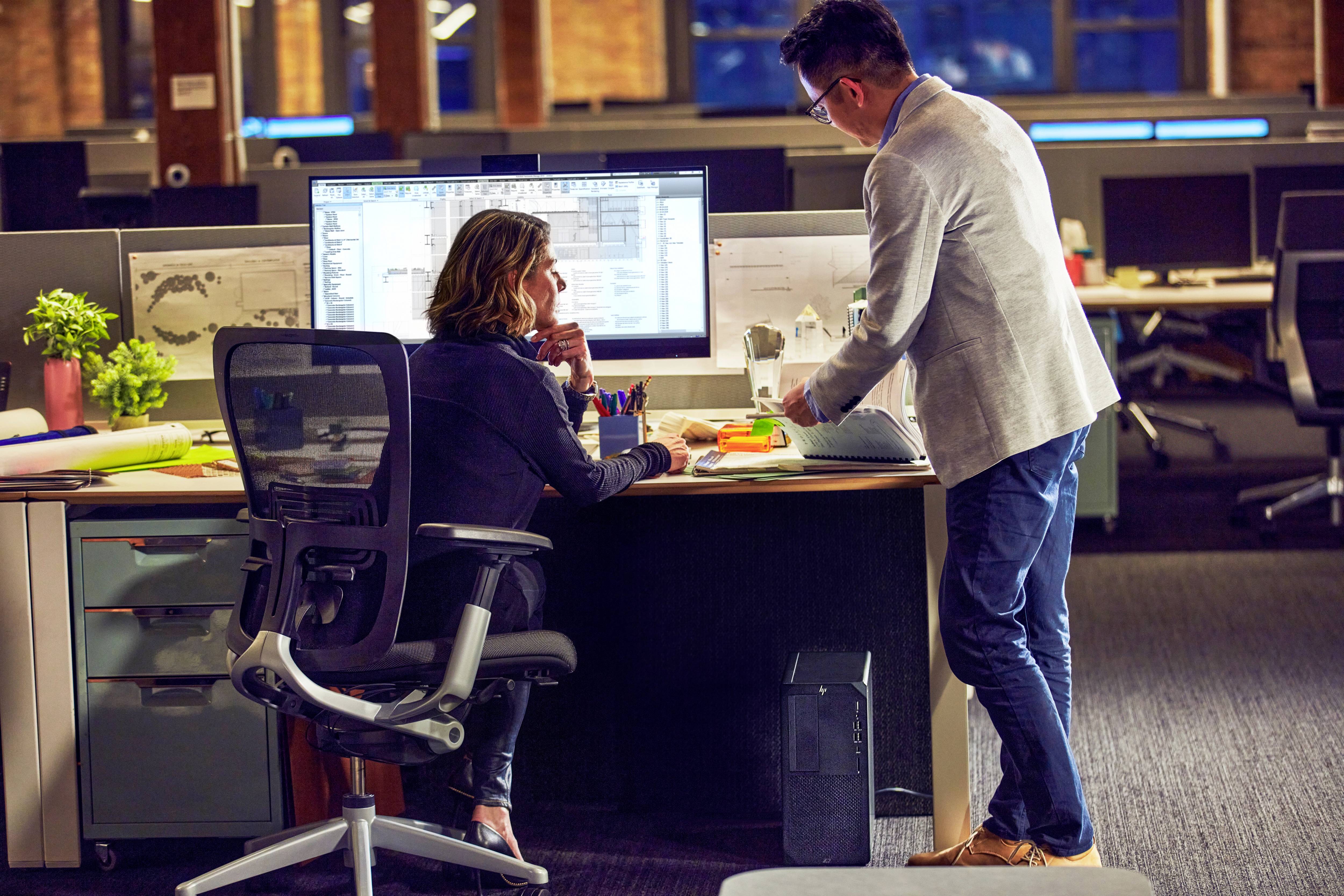Een vrouw zit aan een bureau en werkt op een computer, terwijl een man naast haar staat en naar het scherm wijst. Kantoor op de achtergrond.