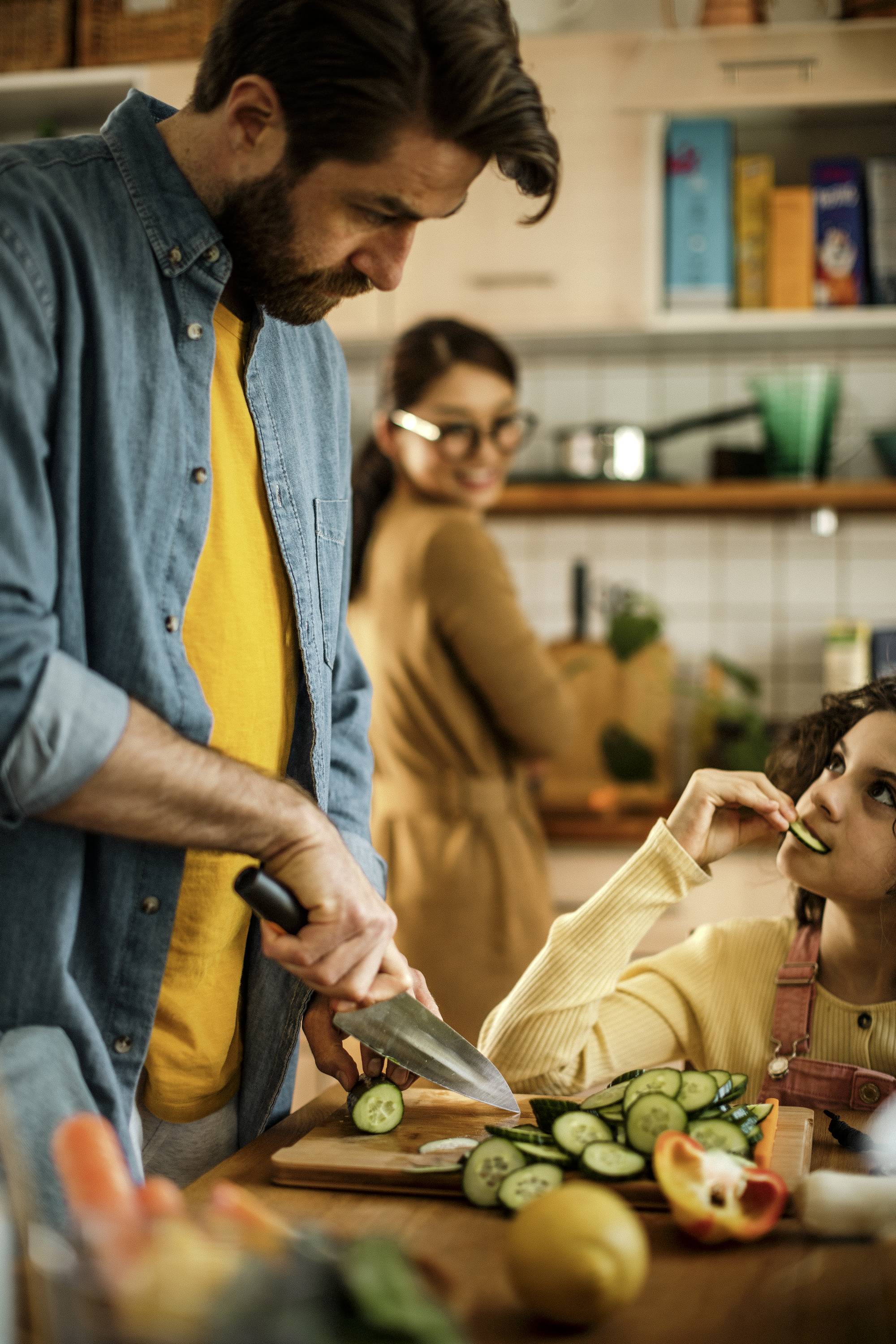 Een man snijdt groente in een keuken, terwijl een kind een stuk komkommer eet en een vrouw op de achtergrond bij het aanrecht staat.