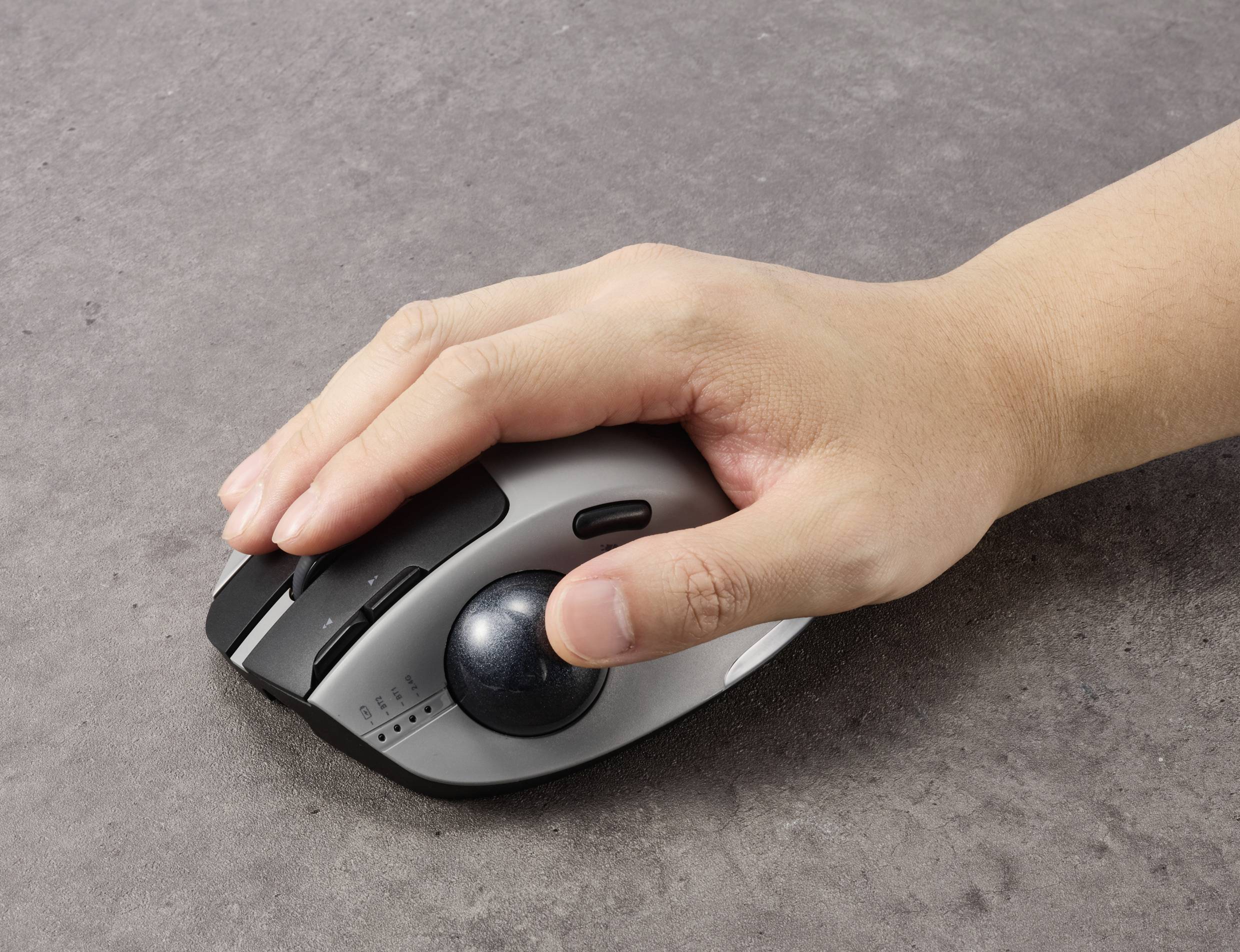 Een hand houdt een ergonomische muis met een duim-trackball op een grijze ondergrond.