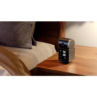 Een moderne tafelklok met digitale weergave staat op een nachtkastje naast een bed. Hij toont de tijd 10:28.
