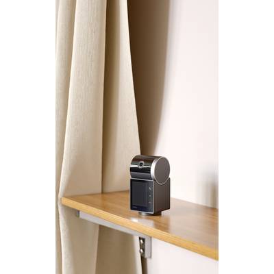 Een klein, modern apparaat met een ronde kop en display staat op een plank voor een beige gordijn.