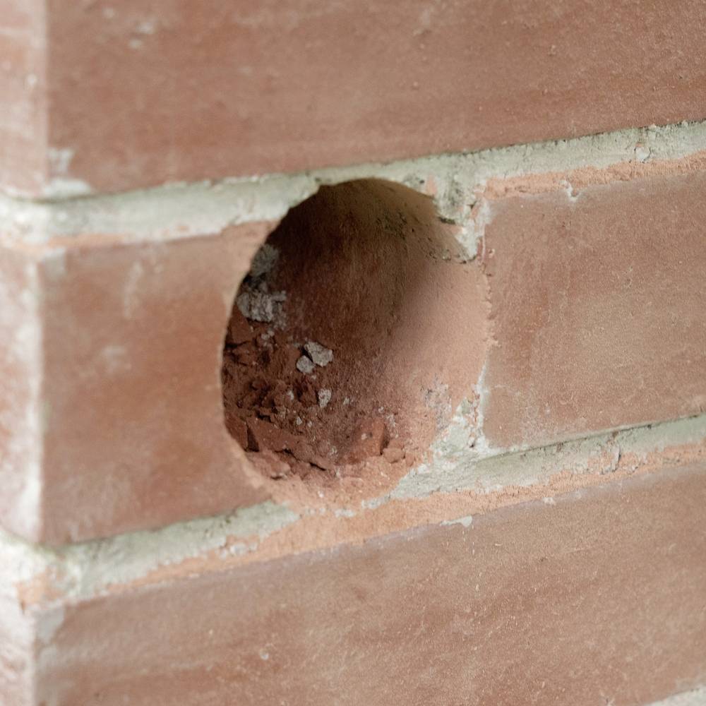 Bakstenen muur met een cirkelvormig gat dat vergruisde puin binnenin toont, wat duidt op beschadiging of voorbereiding voor een installatie.