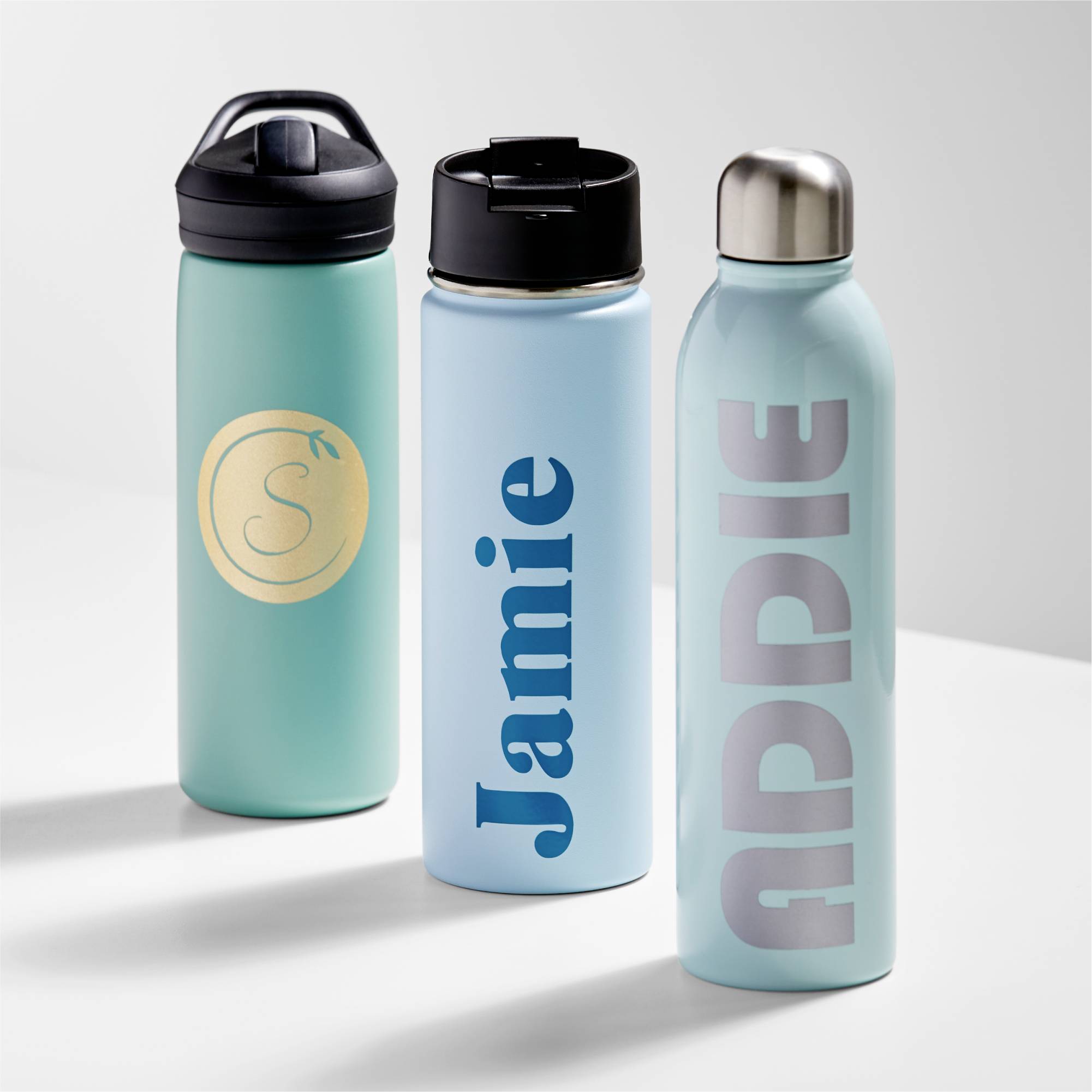 Drie waterflessen naast elkaar op een tafel. Eén heeft een 'S' in een gouden cirkel, de andere dragen de namen 'Jamie' en 'ADDIE'.