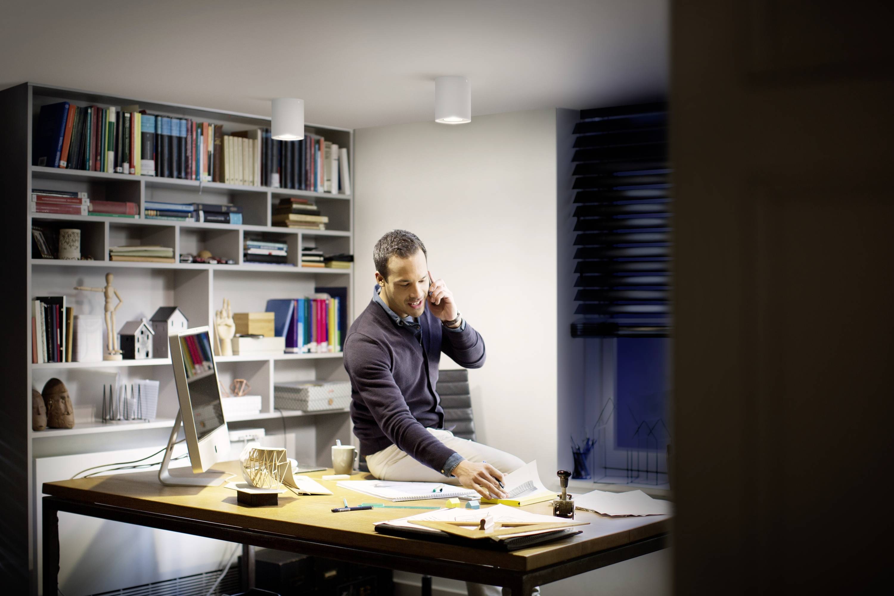 Een man zit aan een bureau in een modern kantoor, belt aan de telefoon en controleert documenten. Boekenplanken op de achtergrond.