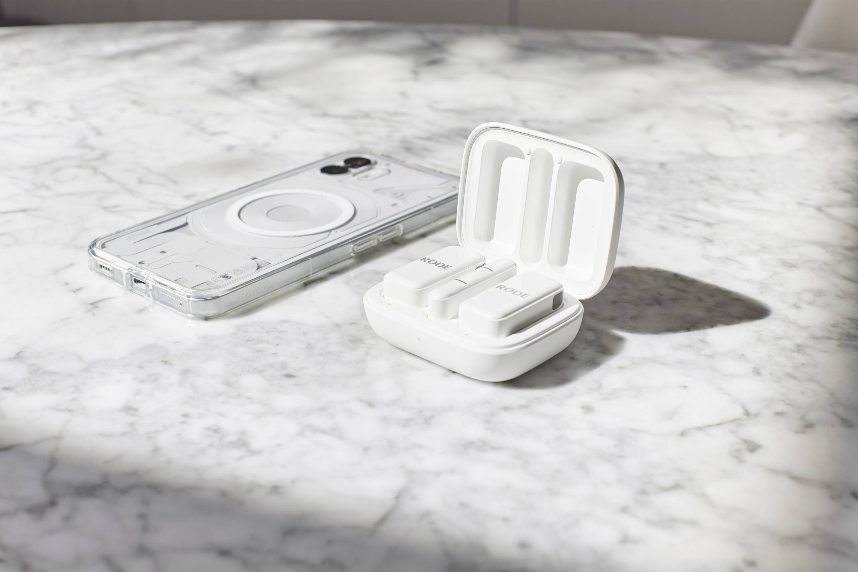 Transparante telefoonhoes en open etui met twee witte draadloze in-ear oordopjes op een marmeren tafel in zonlicht.