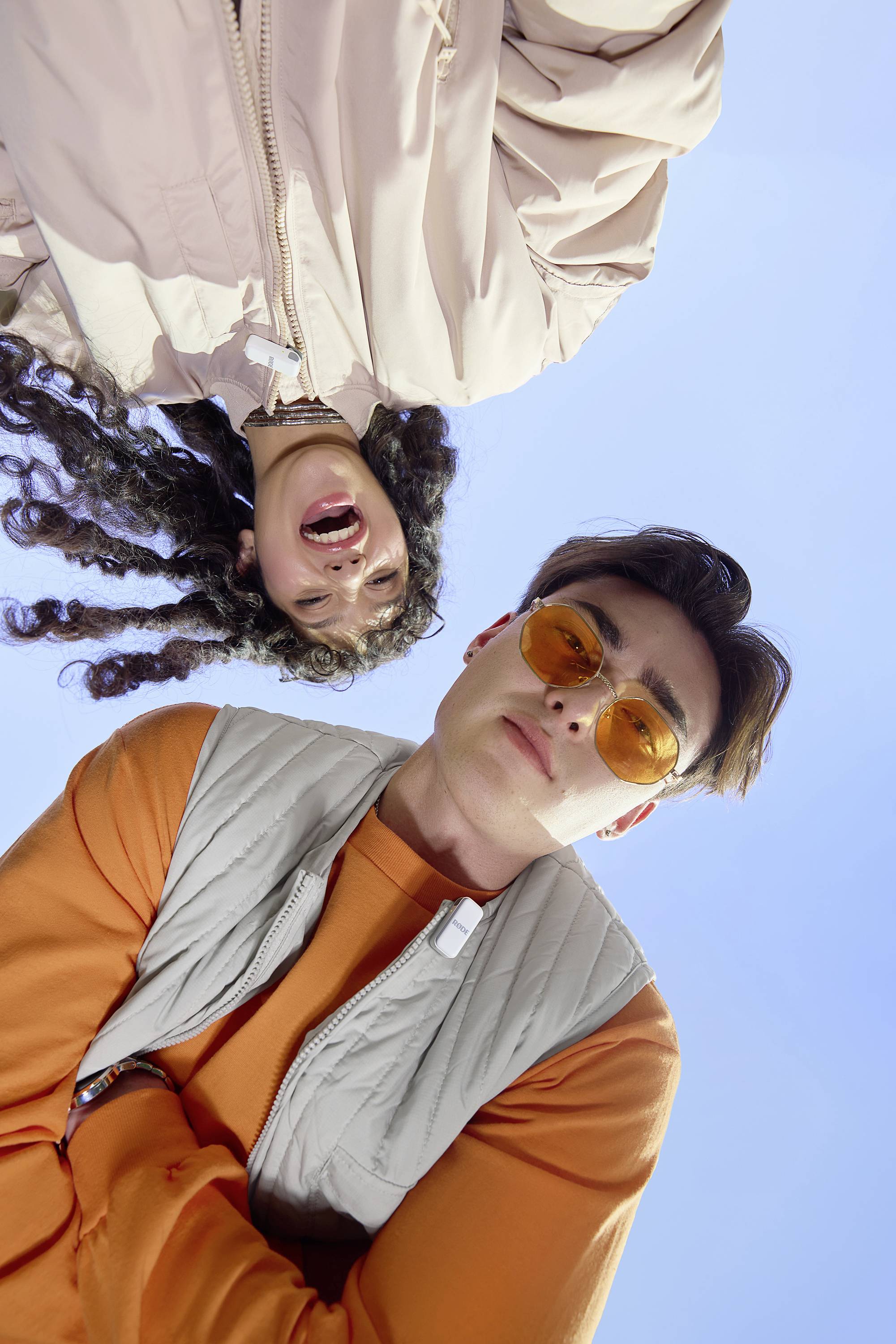 Een man met een oranje zonnebril en een lachende vrouw met krullend haar kijken in de camera in de buitenlucht.