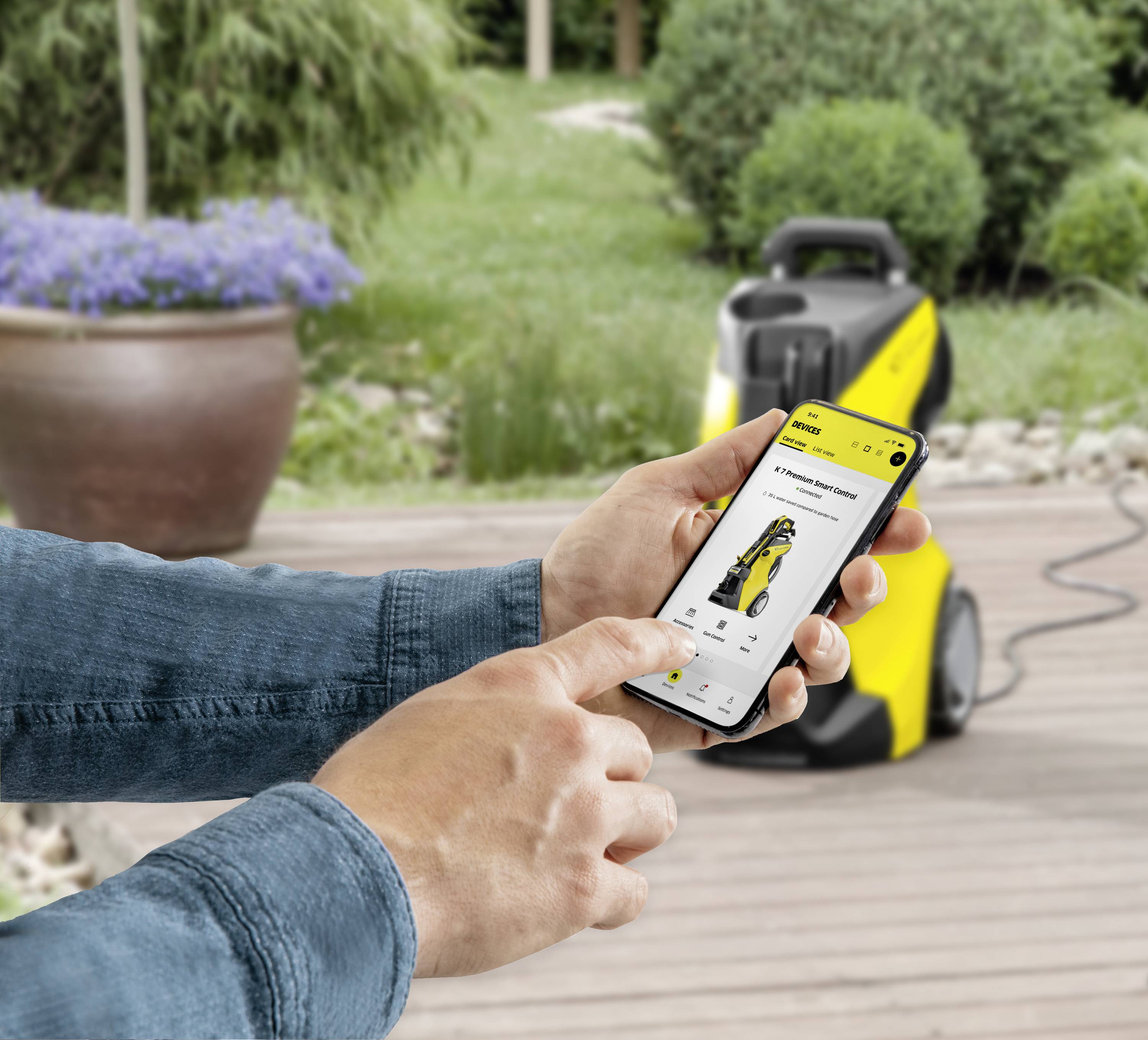Een smartphone toont de besturing van een gele reinigingsmachine in de tuin. Een persoon bedient de app, terwijl de machine op de achtergrond staat.