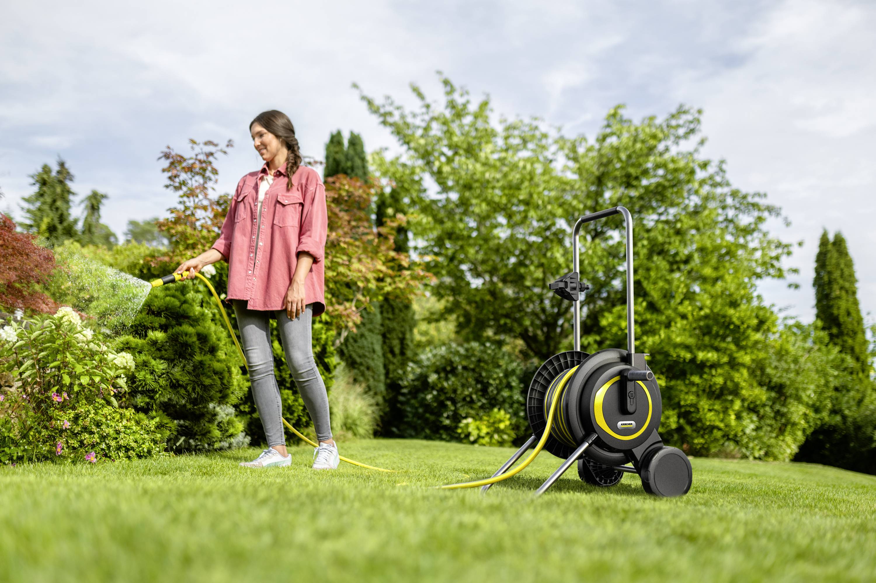 Een vrouw geeft met een tuinslang de tuin water. Op de achtergrond zijn groene struiken en bomen zichtbaar. Naast haar staat een slanghaspel.