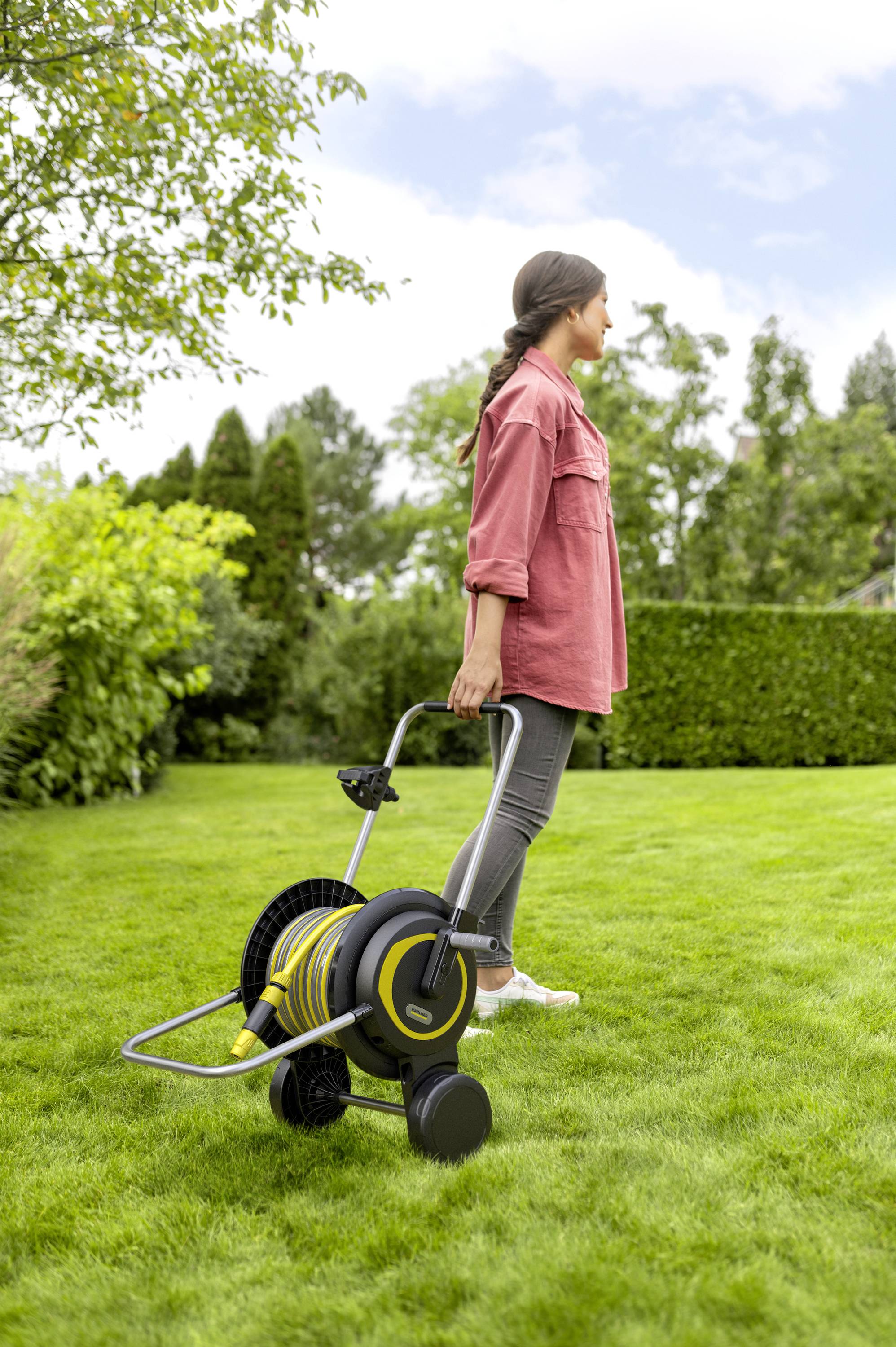 Een persoon trekt een tuinslangwagen over een groen gazon in een tuin. Op de achtergrond zijn bomen en struiken.
