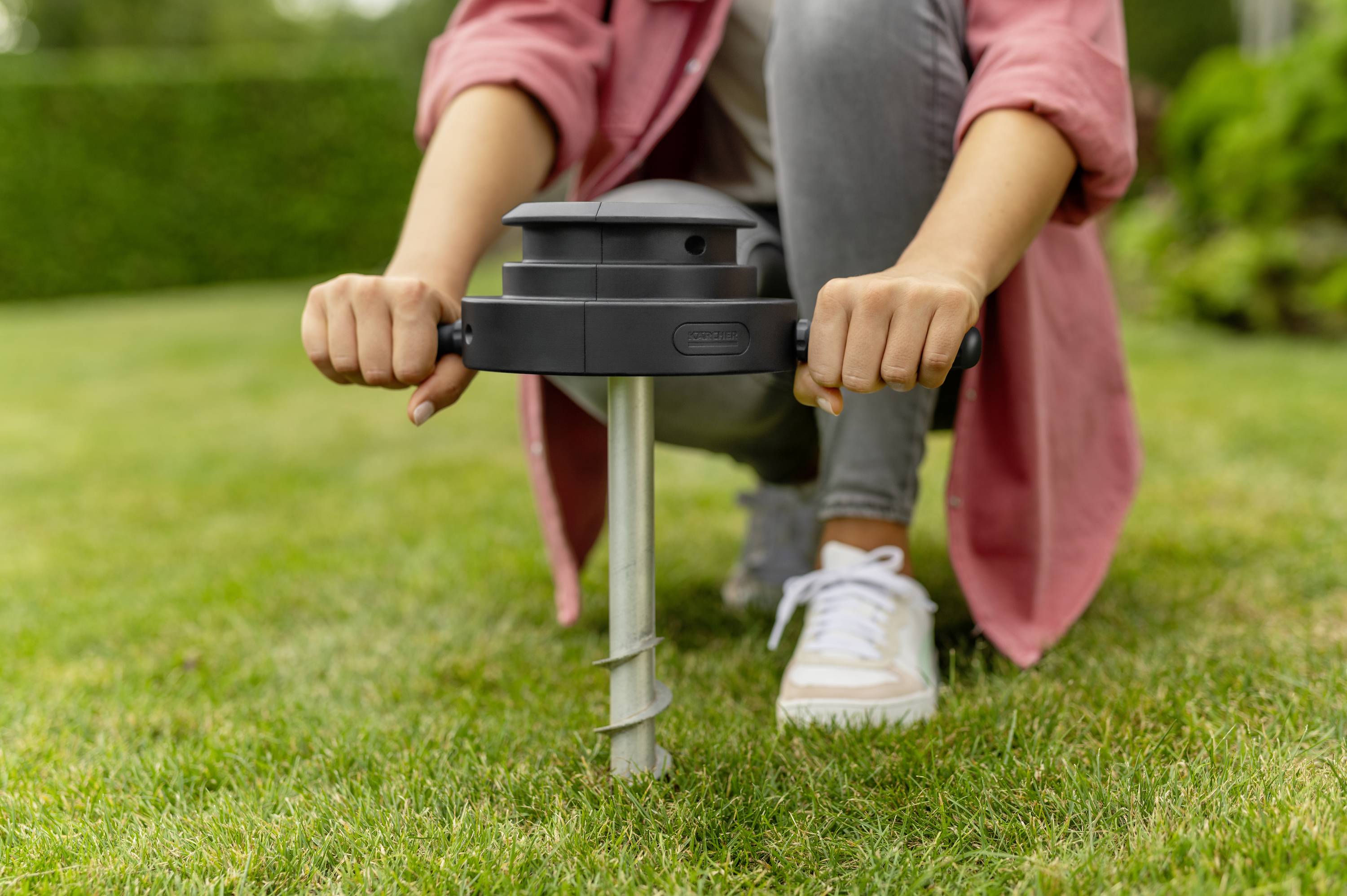 Een persoon installeert een apparaat in het gazon met een handgreep en een schroef. Ze draagt sneakers en een rode jas. Op de achtergrond is een tuin zichtbaar.