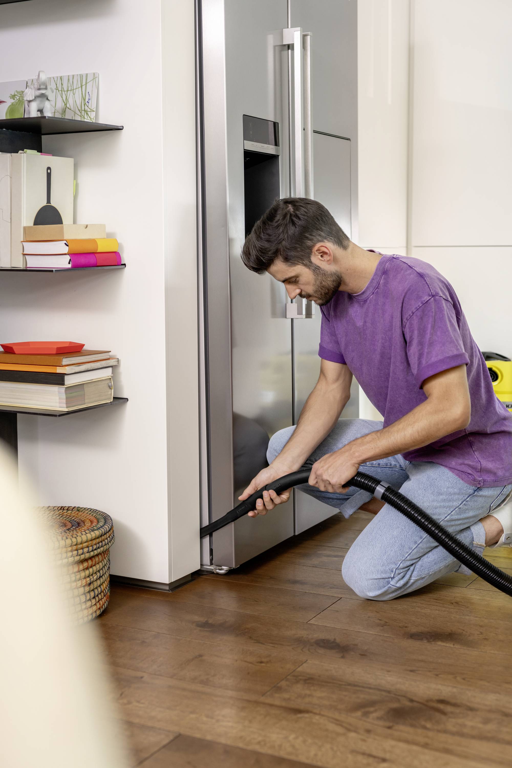Een man knielt op een houten vloer en zuigt met een stofzuiger de hoek van een moderne keuken. Op de achtergrond zijn boekenplanken te zien.