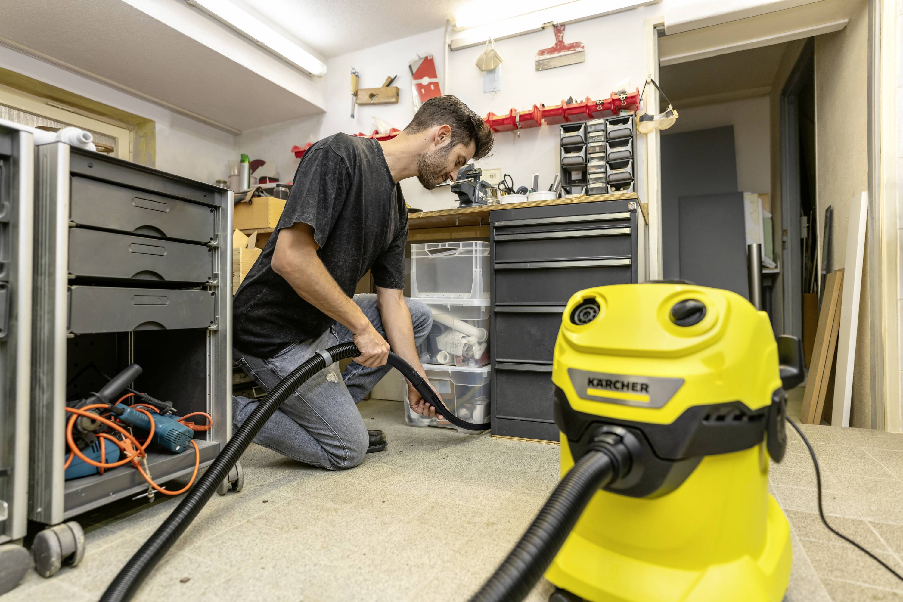 Een man knielt in een werkplaats en gebruikt een gele stofzuiger om de vloer schoon te maken. Op de achtergrond zijn gereedschappen te zien.