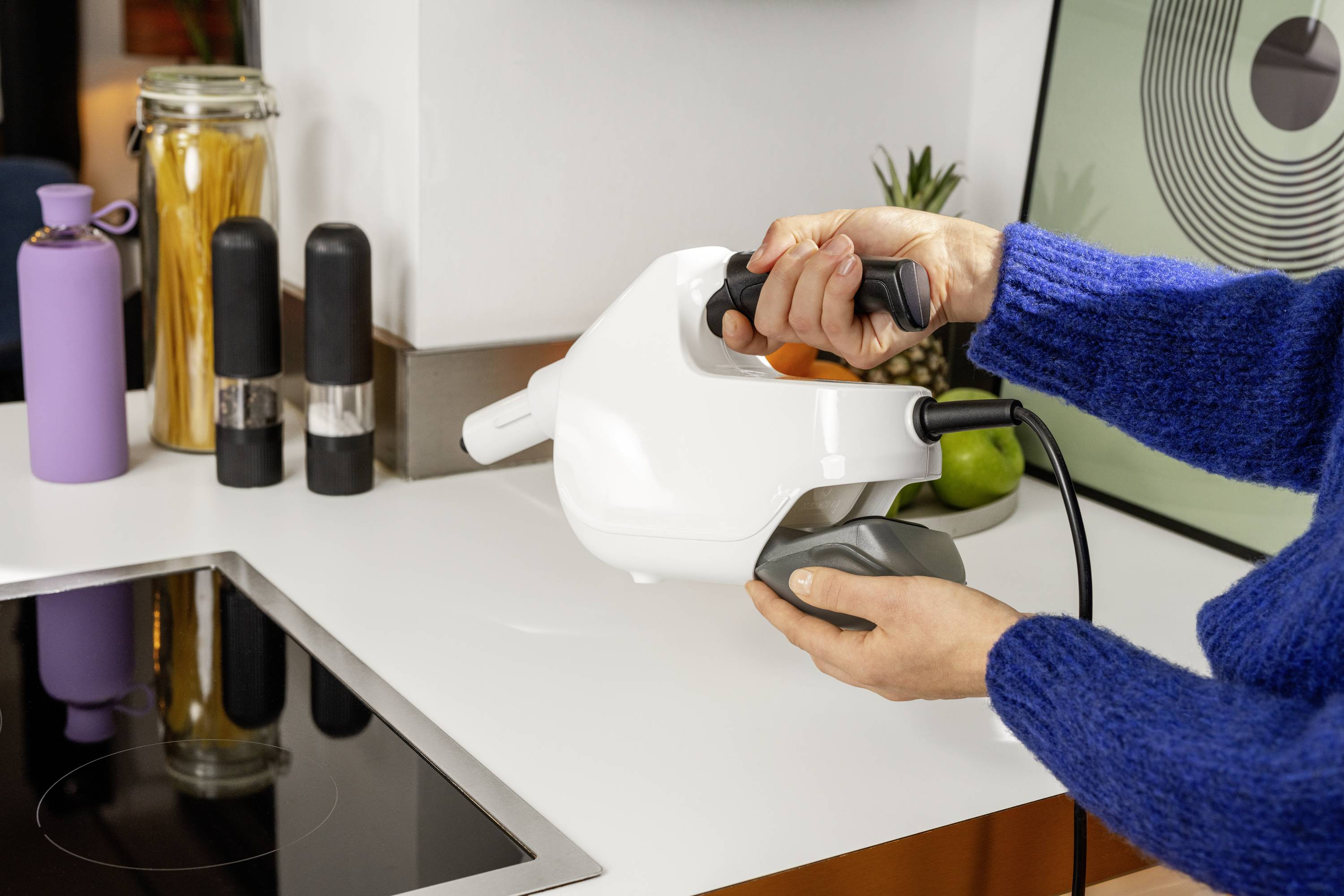Een persoon houdt een witte handstofzuiger boven een wit werkblad in een moderne keuken.