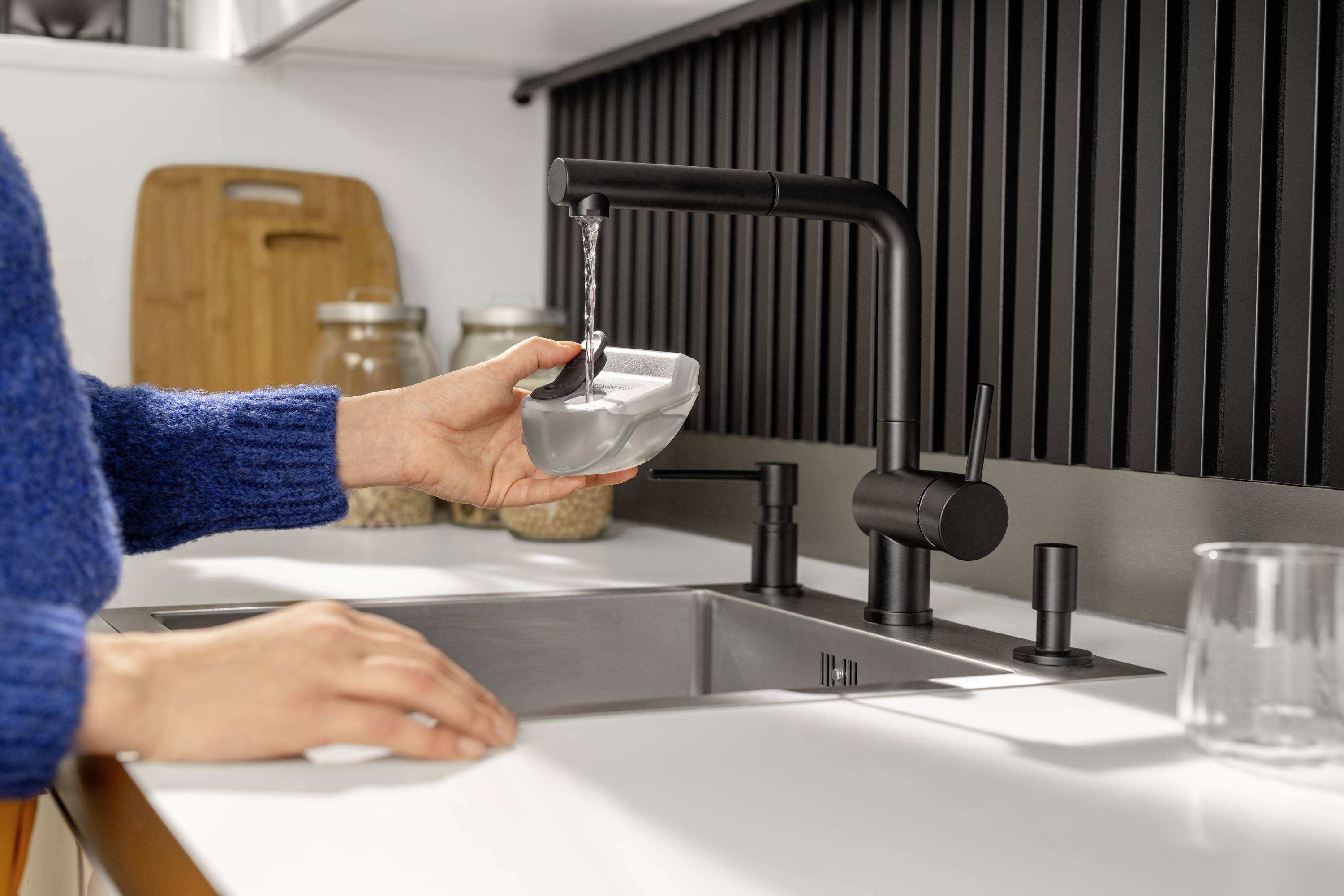 Een persoon vult een grijze kom met water uit een zwarte kraan in een moderne keuken. Op de achtergrond staan voorraadpotten.