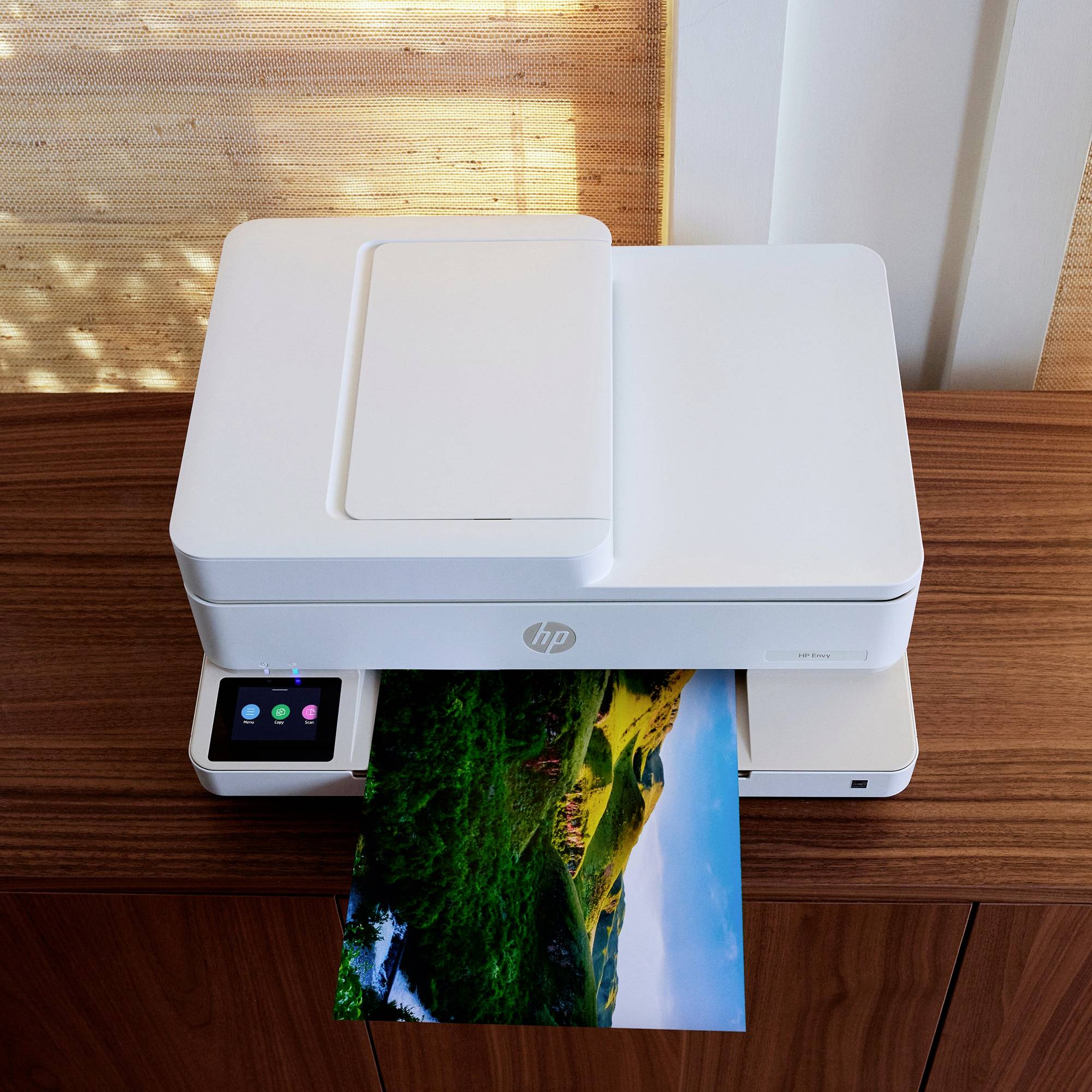 Een witte printer op een houten tafel drukt een foto af met een landschap in levendige groentinten. Links bevindt zich een bedieningspaneel.
