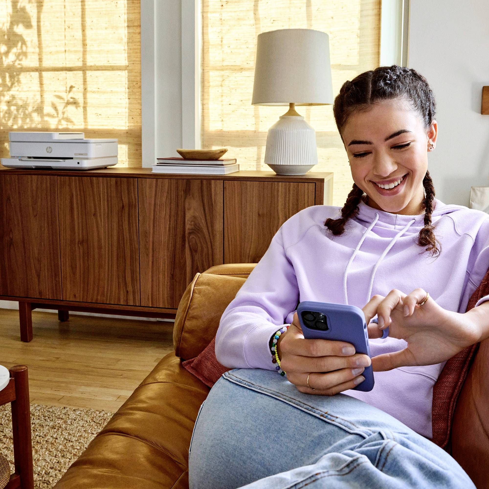 Een lachende persoon zit op een bank en gebruikt een smartphone, met op de achtergrond een printer en planten in een woonkamer.