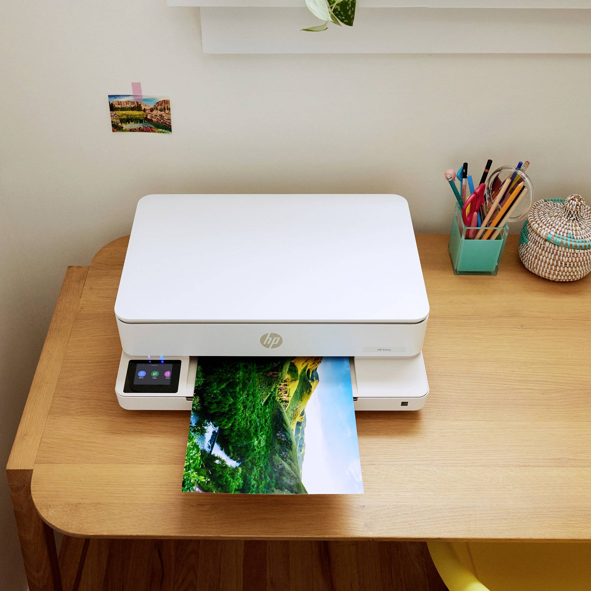 Een printer staat op een bureau en drukt een foto af met een landschapstafereel. Naast de printer staan pennen in een houder.