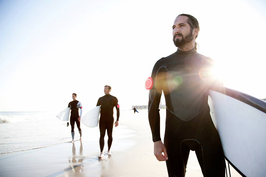 Een man in een wetsuit staat met een surfplank op het strand, terwijl twee personen op de achtergrond langs de kustlijn joggen.
