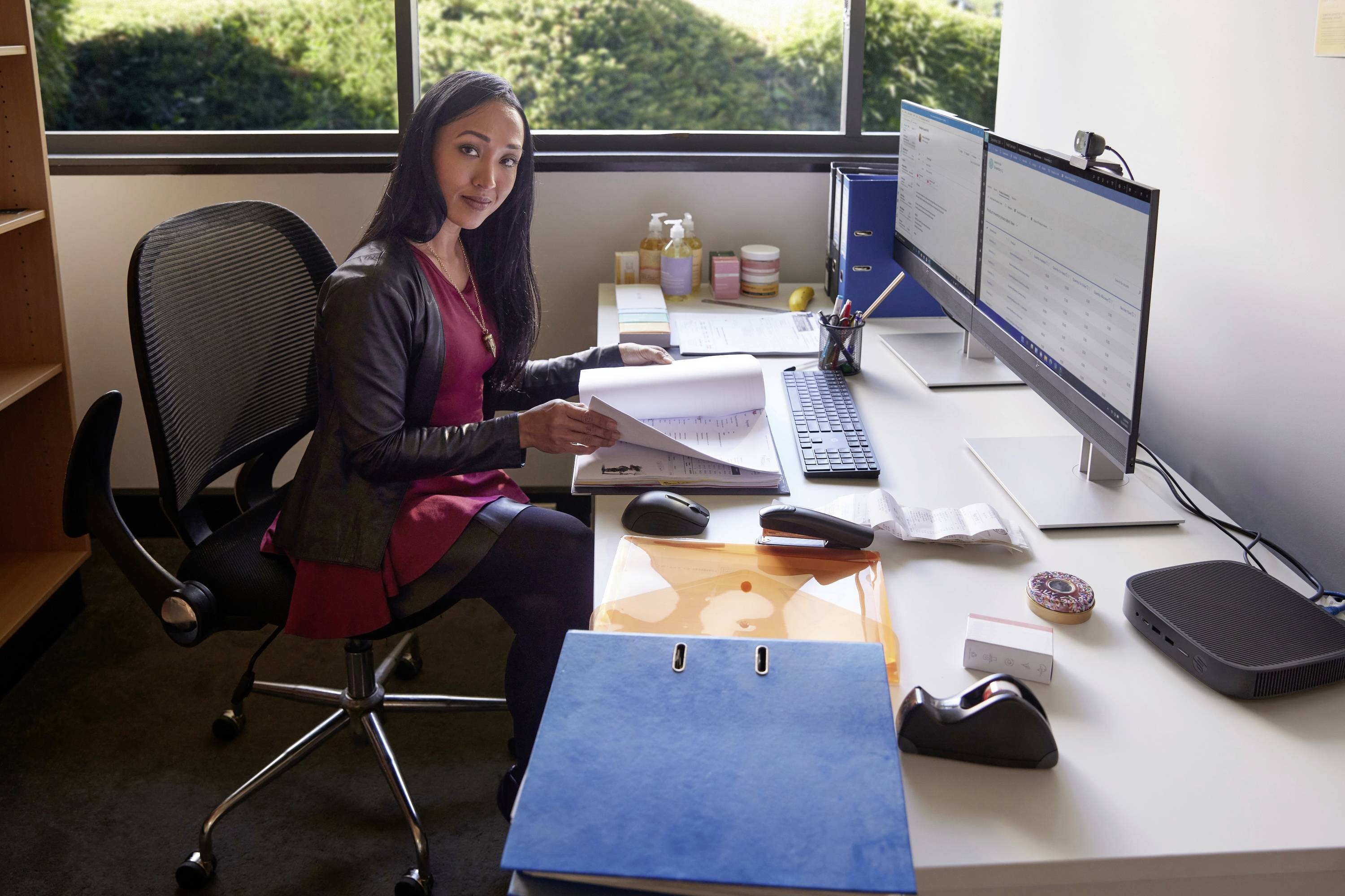 Een vrouw zit in een kantoor aan een bureau, bekijkt documenten op twee monitoren. Op de achtergrond is een raam met uitzicht naar buiten.
