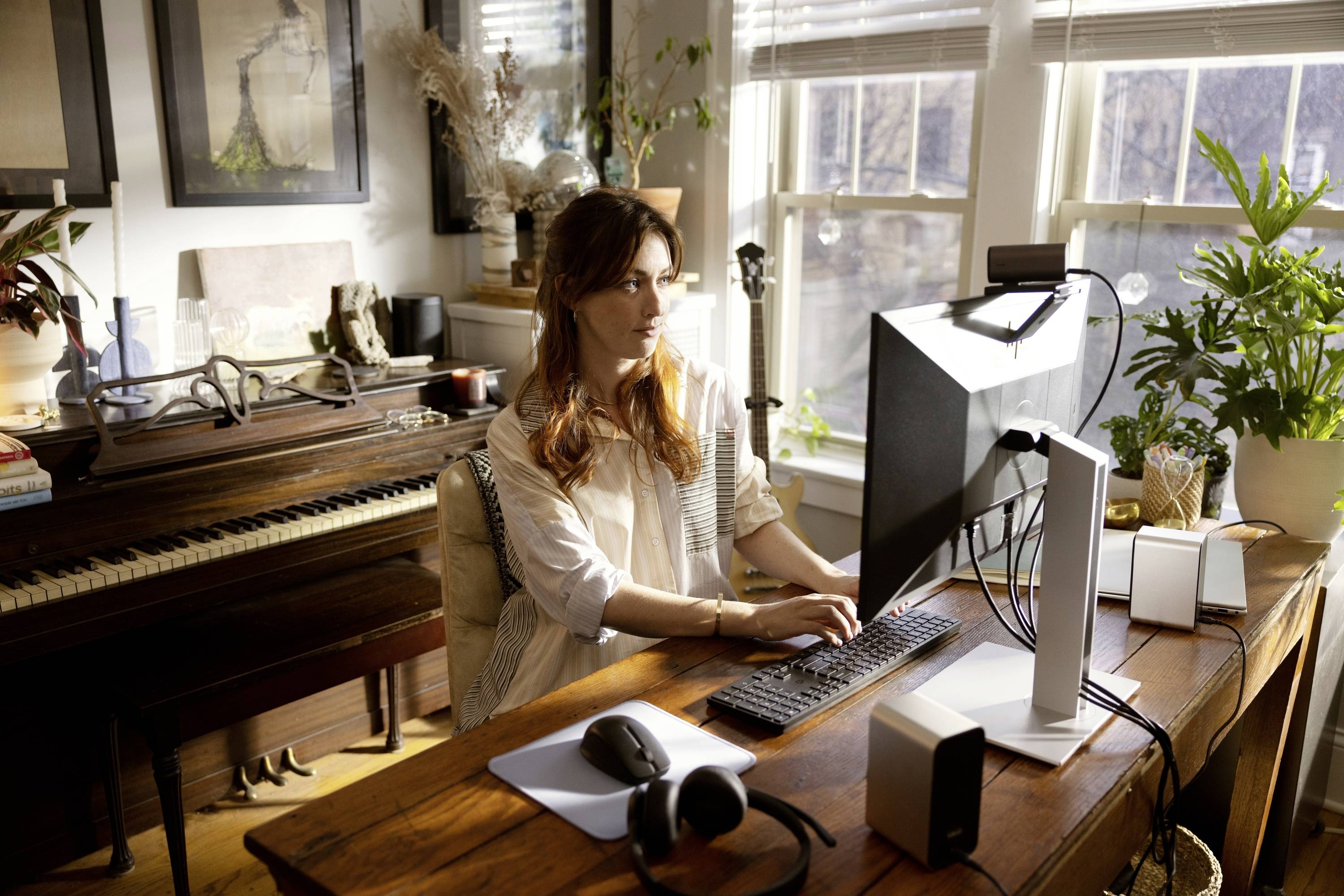 Een vrouw werkt geconcentreerd op de computer in een lichte ruimte met veel planten en een piano op de achtergrond.