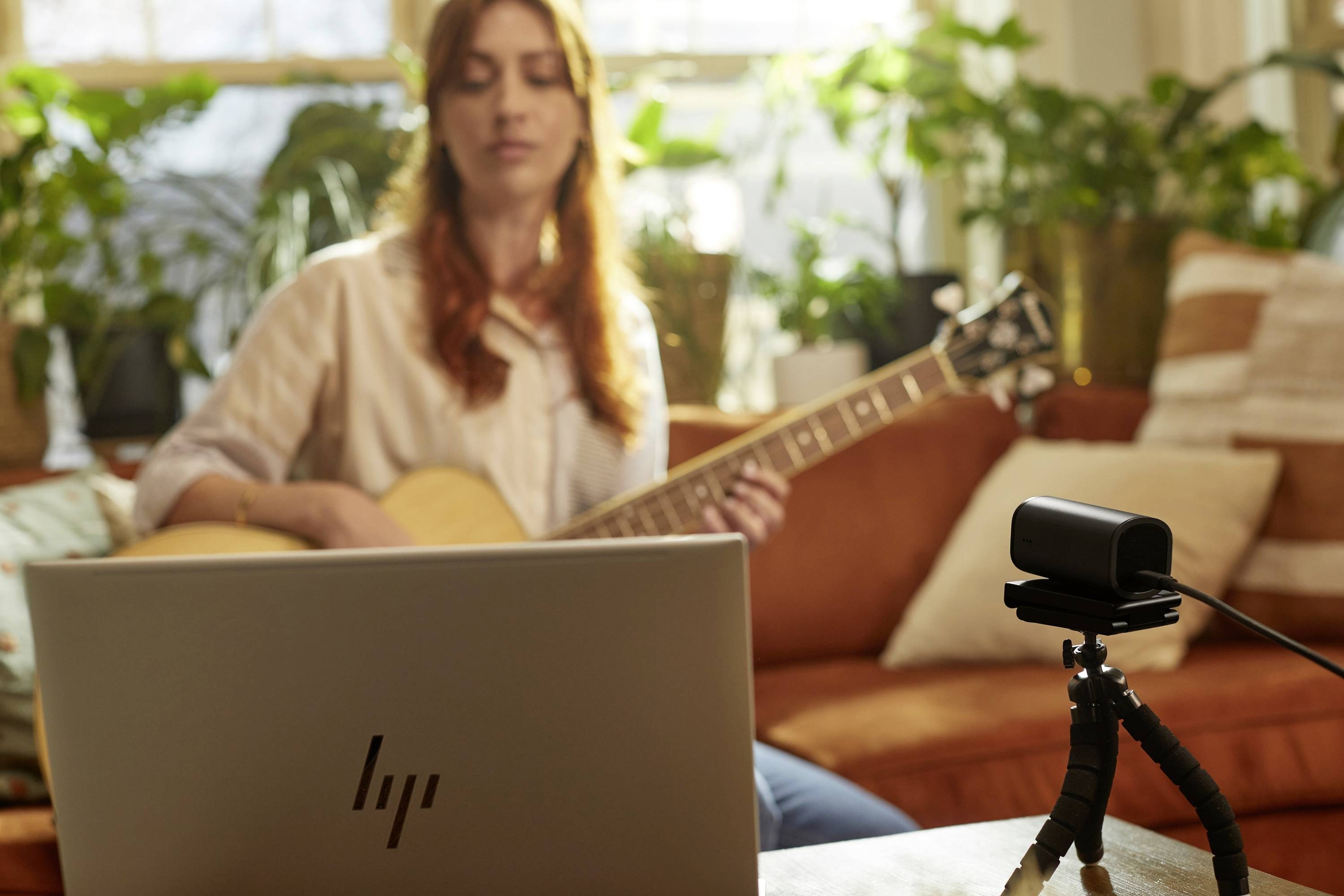 Een vrouw speelt gitaar voor een laptop en een camera. Op de achtergrond zijn planten zichtbaar.