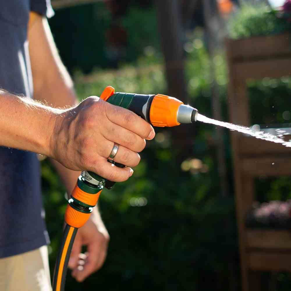 Een persoon besproeit planten met een tuinslang in het zonlicht. De focus ligt op de waterstraal en de hand die de slang vasthoudt.