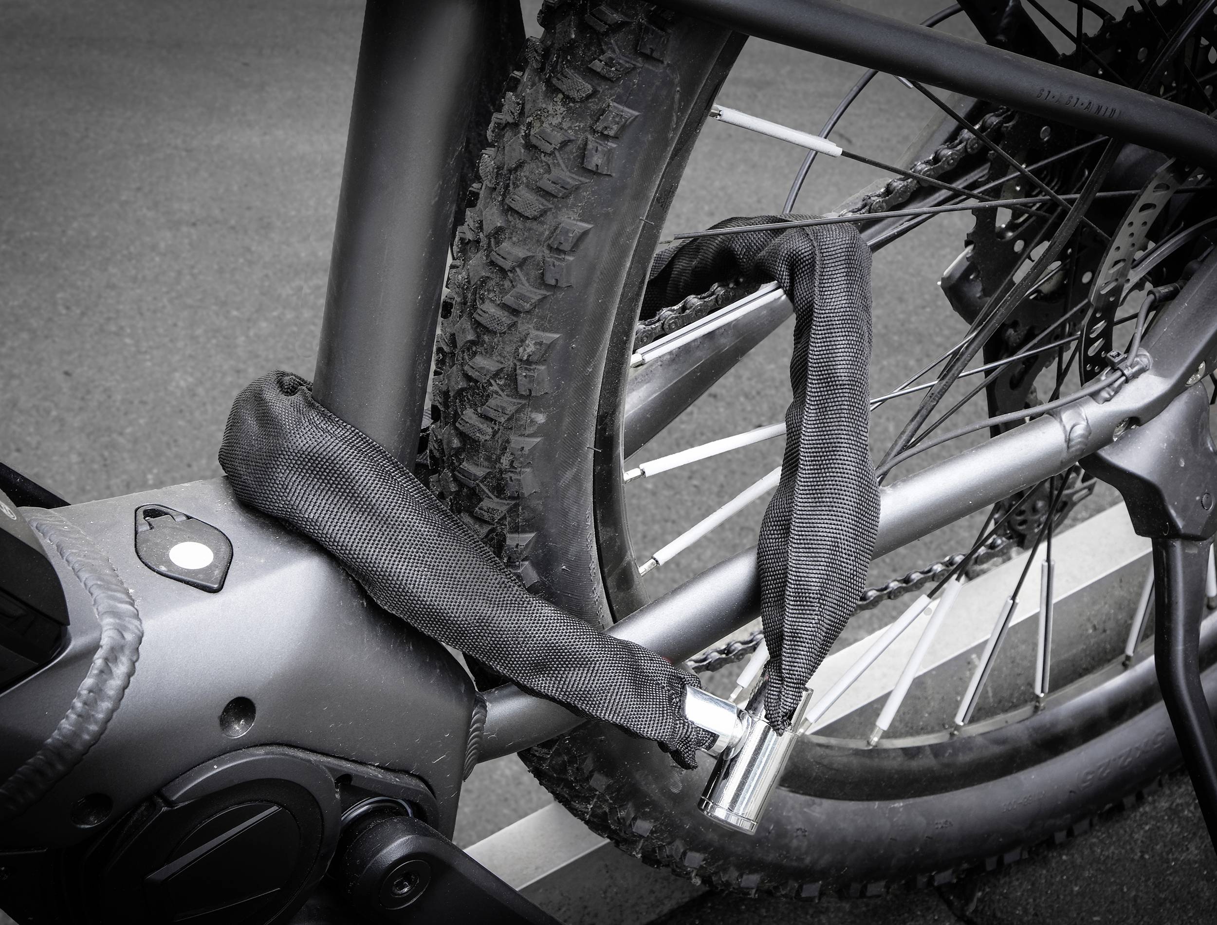 'Close-up van een fietsframe met een kettingslot dat is bevestigd rond het achterwiel, op een geassfalteerd oppervlak.'