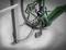 Een groene fiets is met een zwart kabelslot vastgemaakt aan een metalen standaard. De focus ligt op het slot en de achterband.