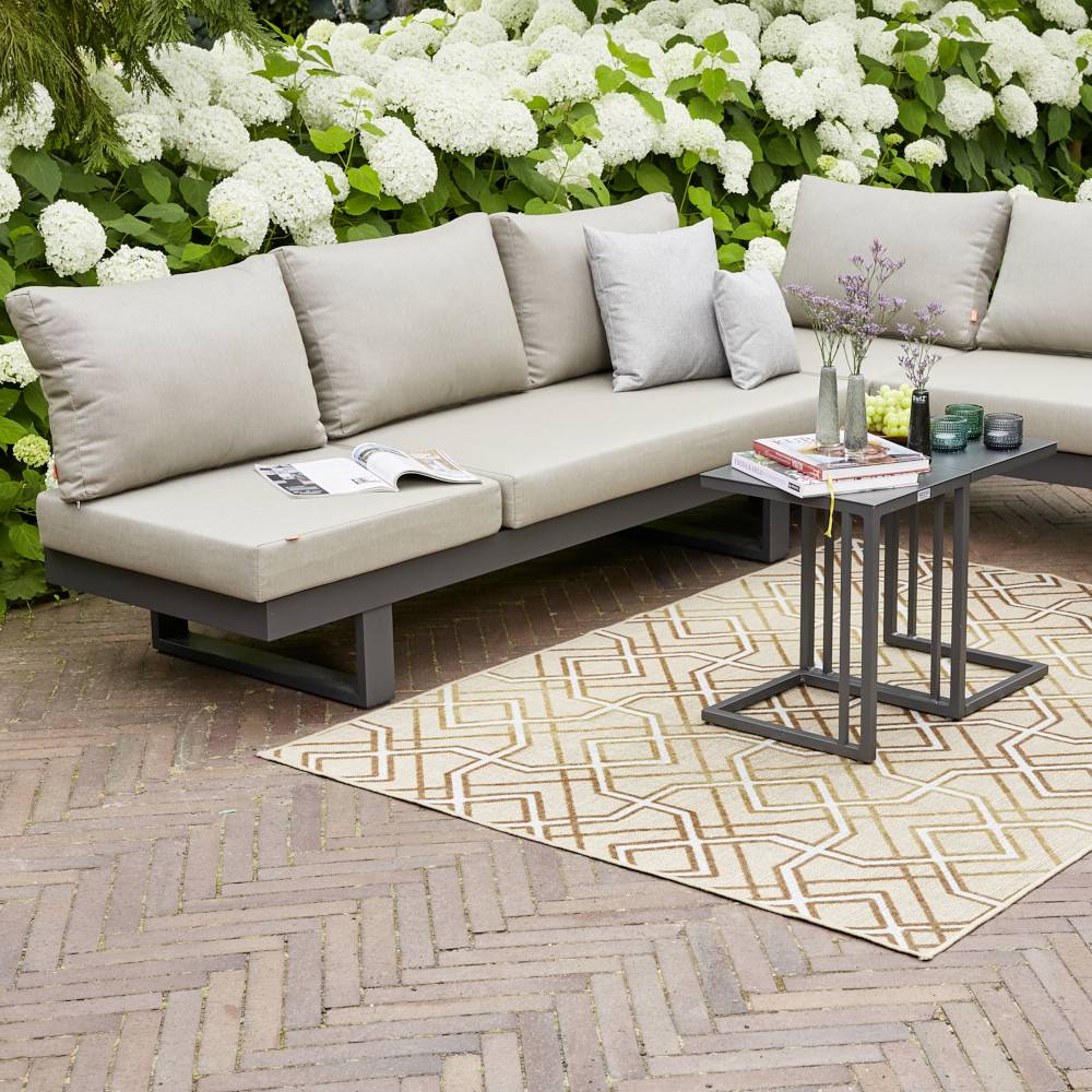 Een grijze hoekbank met kussens staat op een terras, omringd door bloeiende witte hortensia's. Een vloerkleed en een bijzettafel completeren de scène.