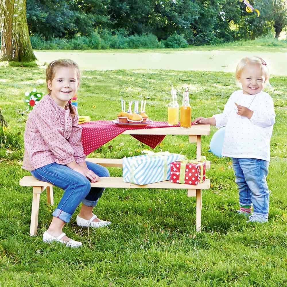 Twee kinderen zitten aan een gedekte picknicktafel buiten. Op tafel staan dranken en cadeaus. Ze lachen en zien er blij uit.