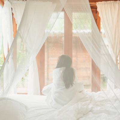 Een vrouw zit op een bed naast een groot raam, waar natuurlijk licht en groene planten doorheen schijnen.