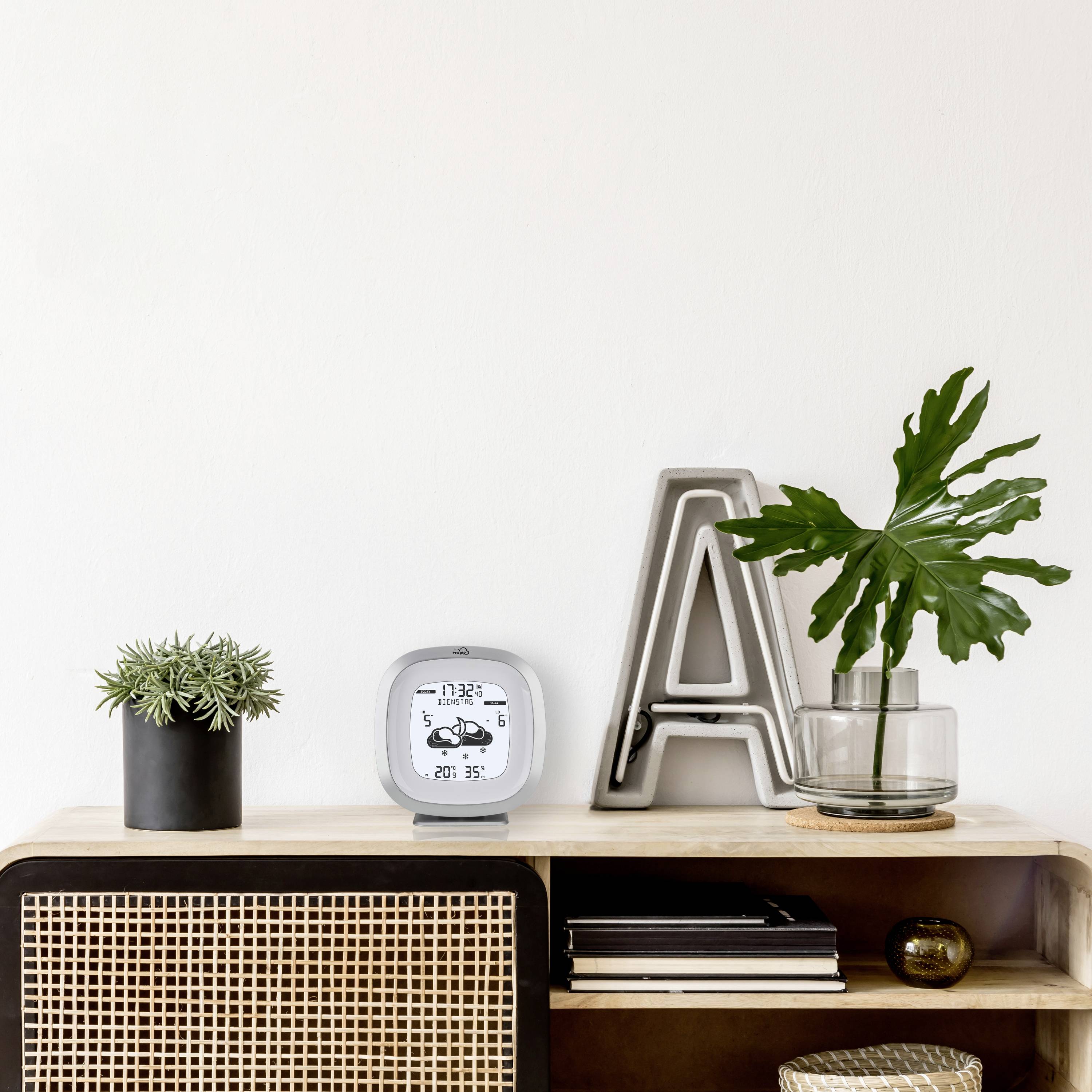 Een rek met een digitale klok, planten en decoratieve letters op een gladde oppervlakte. Op de achtergrond een witte muur.