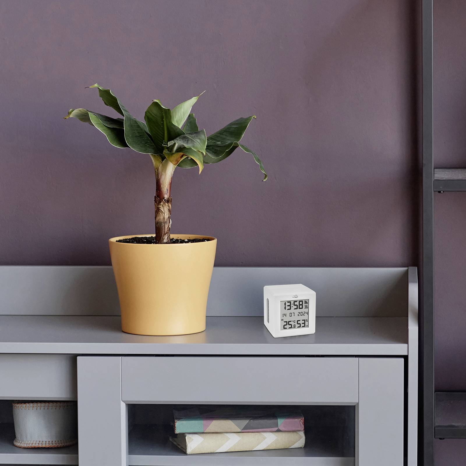 Een groene plant in een gele pot staat op een grijze tafel. Ernaast bevindt zich een kleine, witte digitale wekker.