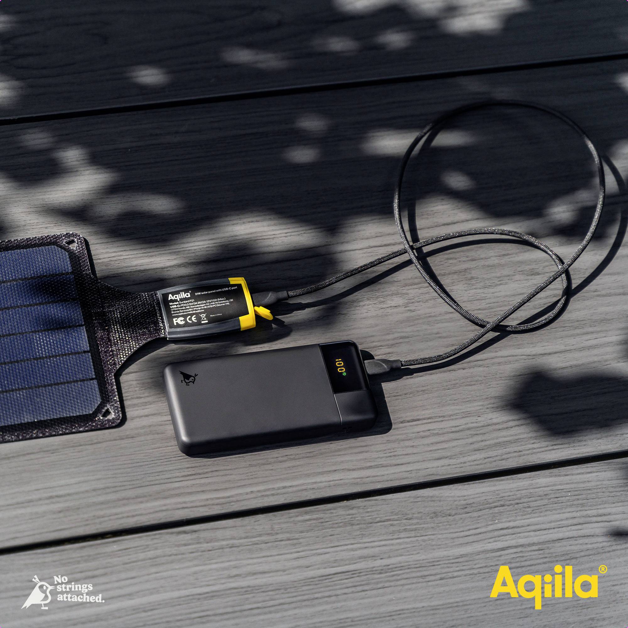 Zonnestroombank laadt een apparaat op via een kabel. Op de powerbank staat '100%'. Schaduwen van bladeren sieren de houten ondergrond.