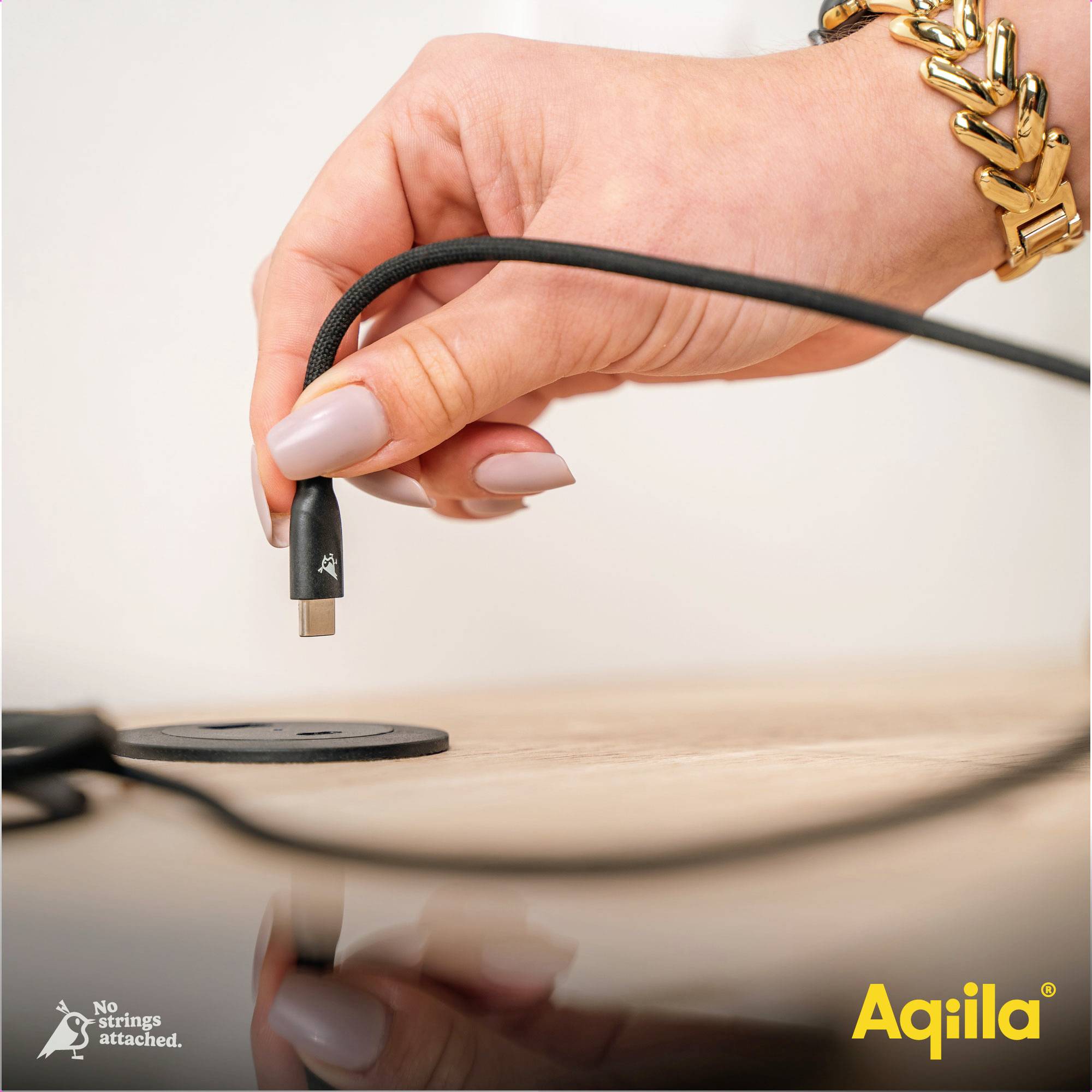 Een hand tilt een magnetische kabel van de tafel, waarop de kabel 'No strings attached.' staat en rechtsonder het logo 'Aqila'.