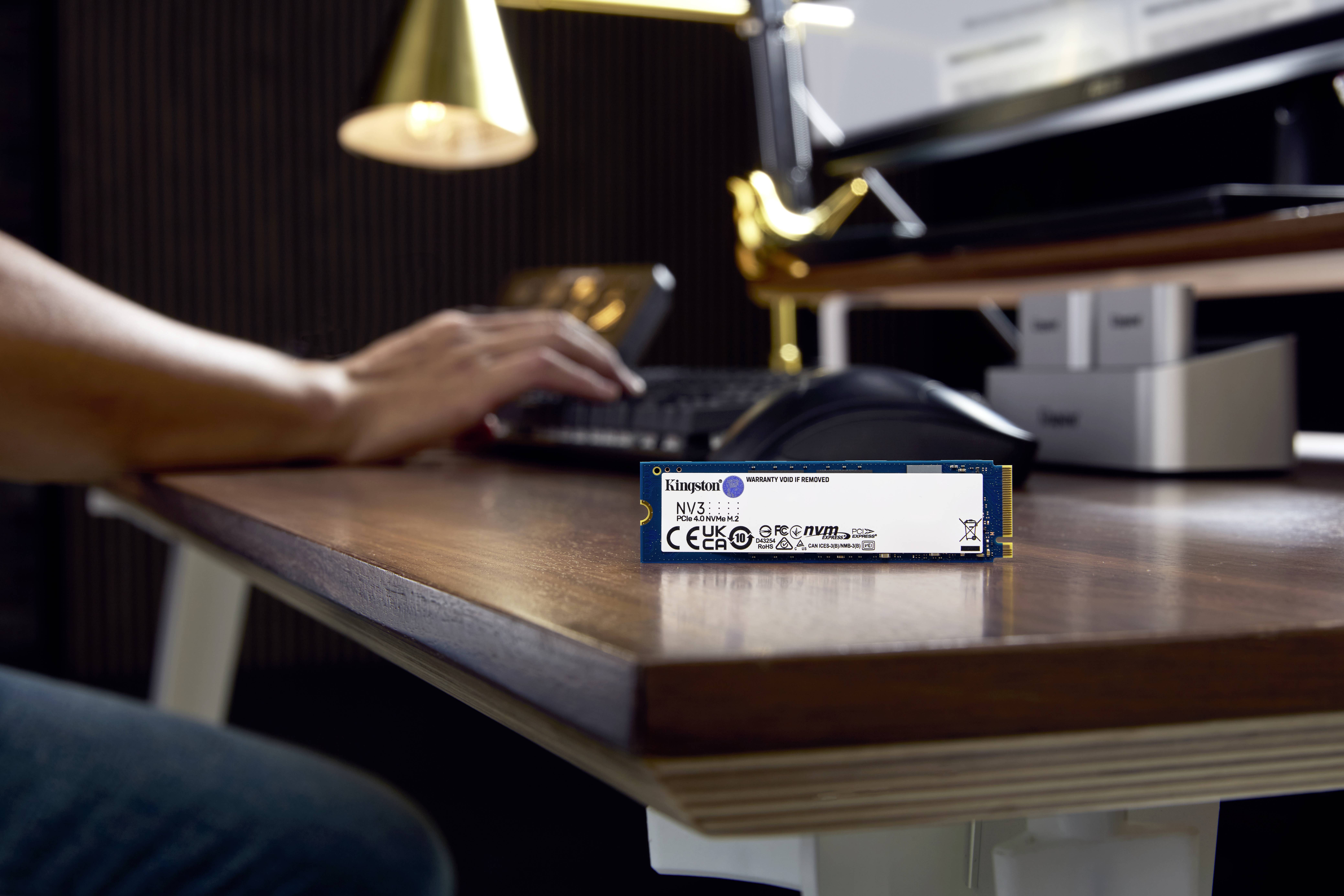 Een M.2 NVMe SSD-schijf ligt op een bureau op de voorgrond. Op de achtergrond typt iemand op een toetsenbord.