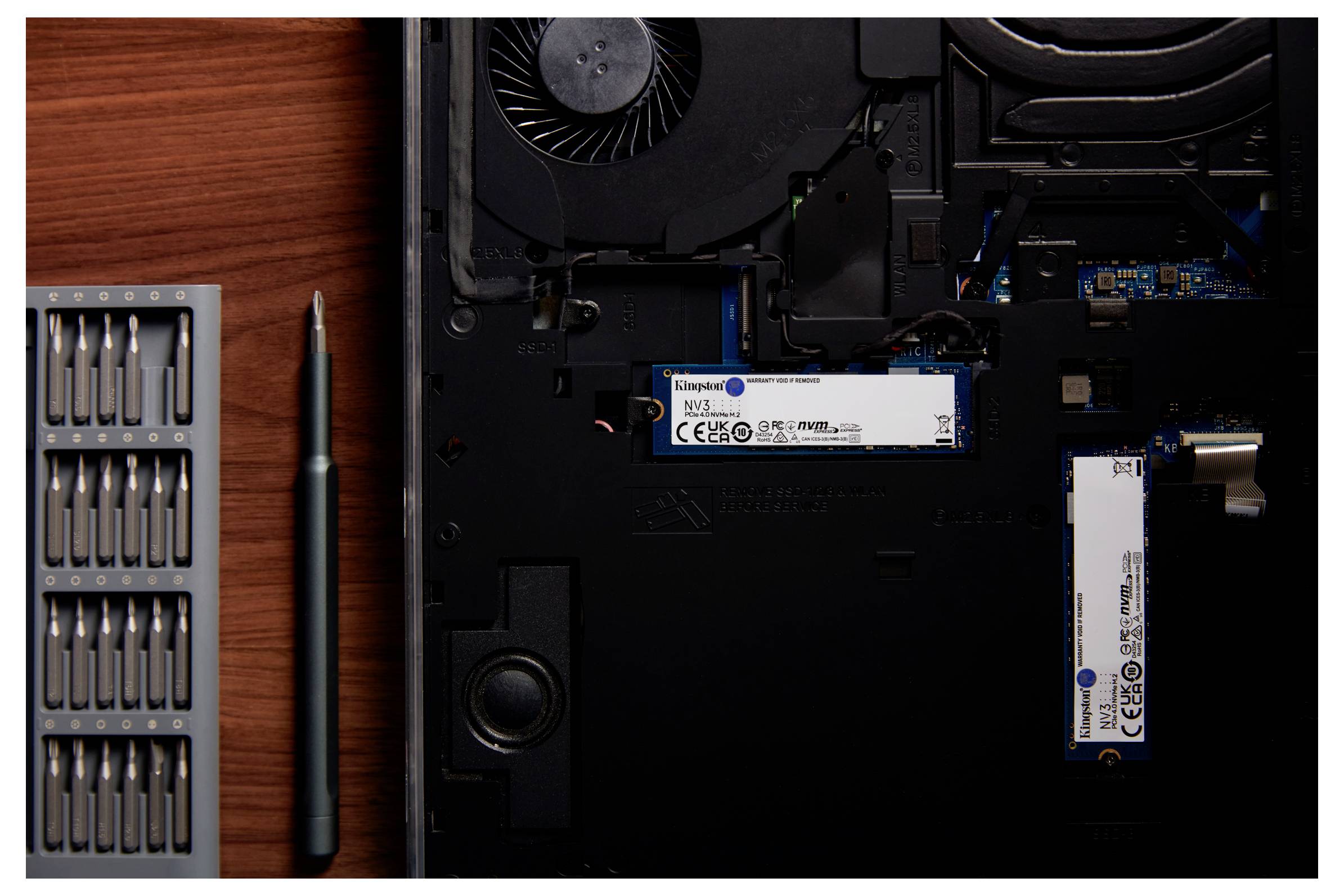 Op de afbeelding is de binnenkant van een laptop te zien met een geopend compartiment dat twee SSD's en een ventilator toont. Ernaast ligt een schroevendreher-set.
