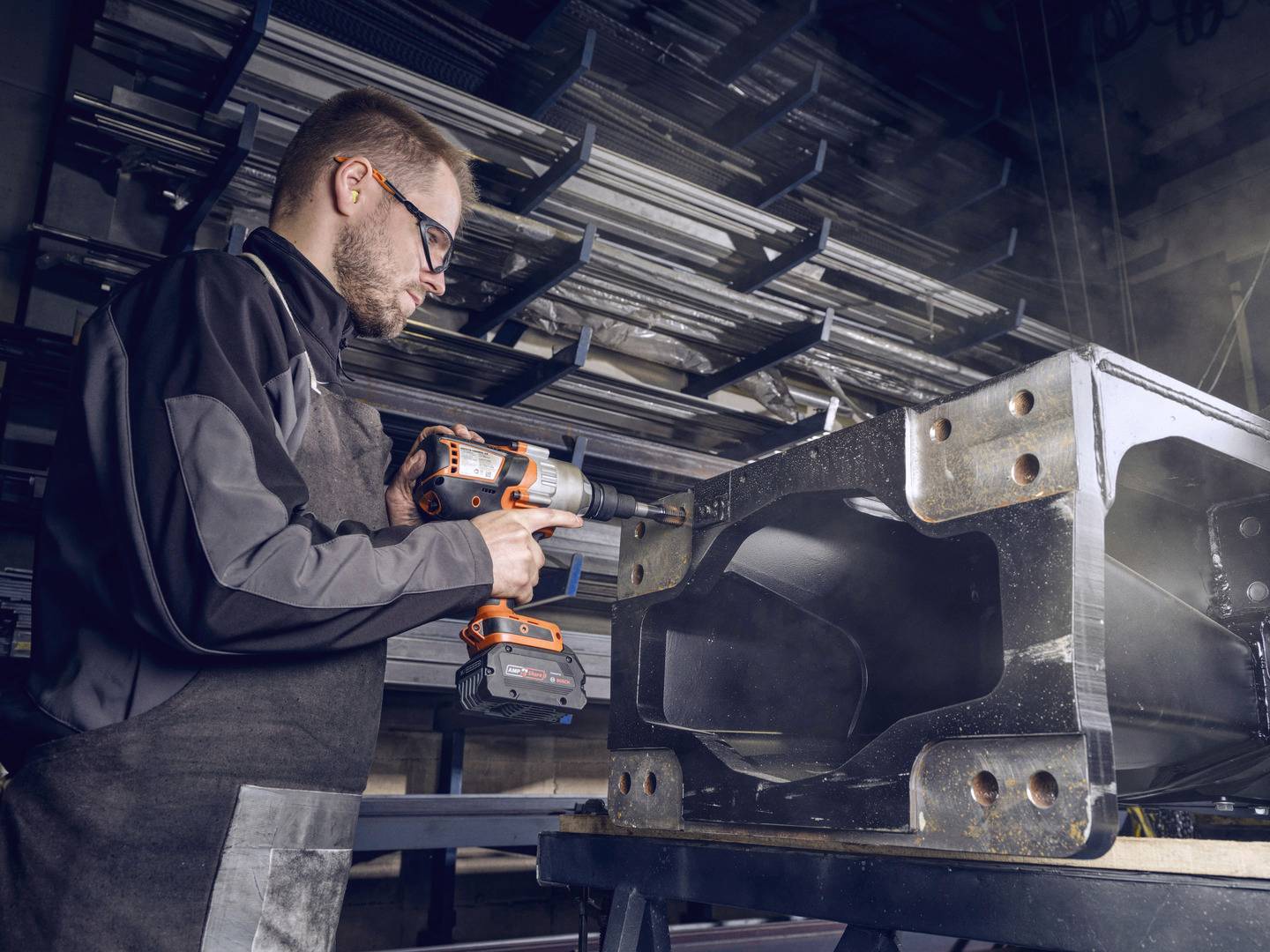 Een man in beschermende kleding gebruikt een boormachine om een groot metalen onderdeel te bewerken in een werkplaats. Op de achtergrond zijn metalen stellingen zichtbaar.