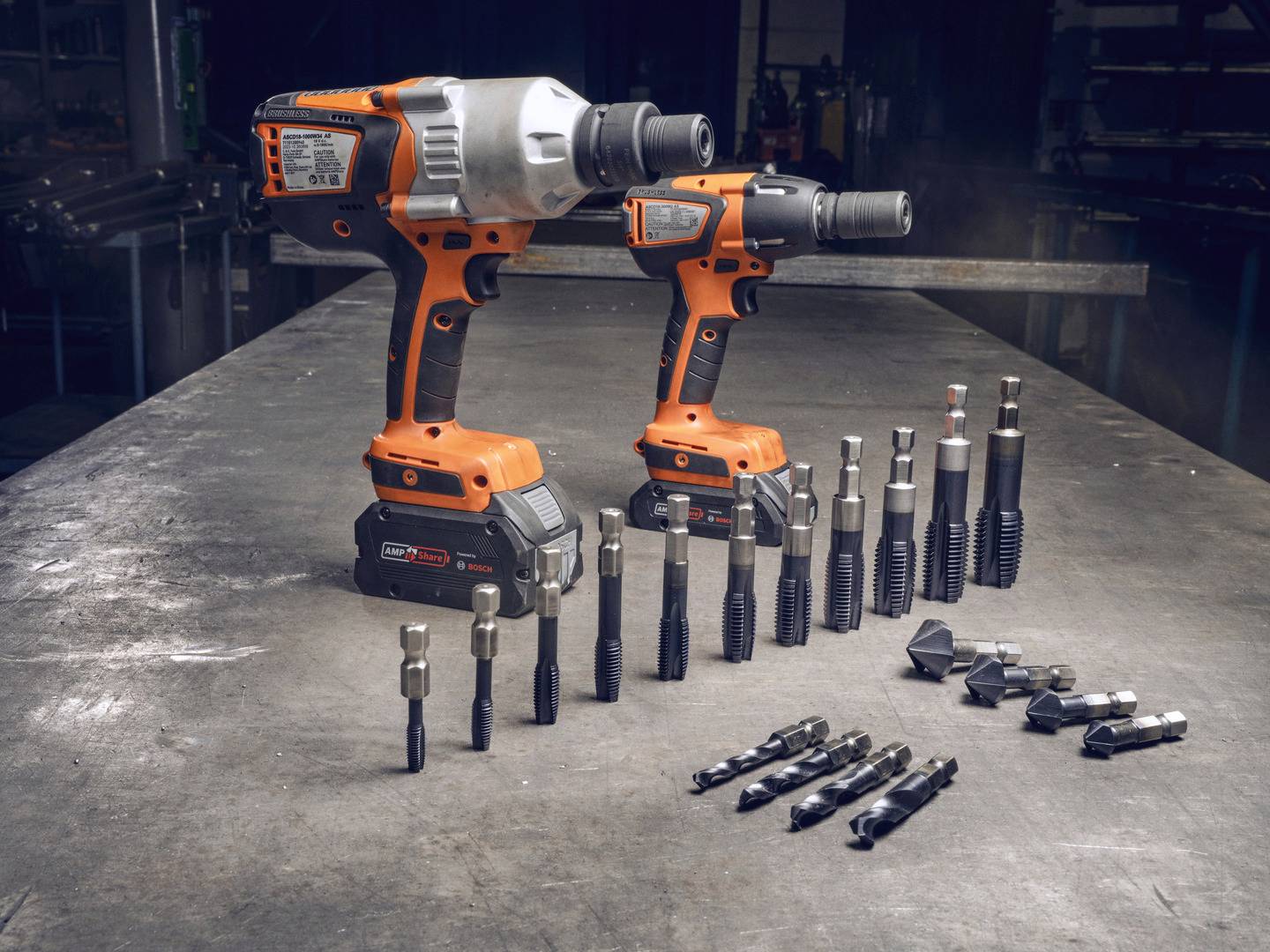 Twee oranje accuschroefmachines met toebehoren op een werkbank. Links staat de grotere schroevendraaier, rechts de kleinere. Verschillende boor- en schroefopzetstukken liggen vooraan gerangschikt.