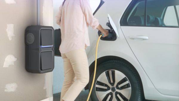 Een persoon laadt een witte elektrische auto op bij een wandoplaadpunt. Ze houdt het laadkabel vast en staat naast de auto.