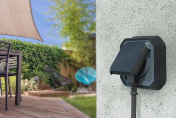Een buitenstopcontact aan een muur van een huis met een ingeplugd kabel. Op de achtergrond een terras met tuinmeubels en veel groen.